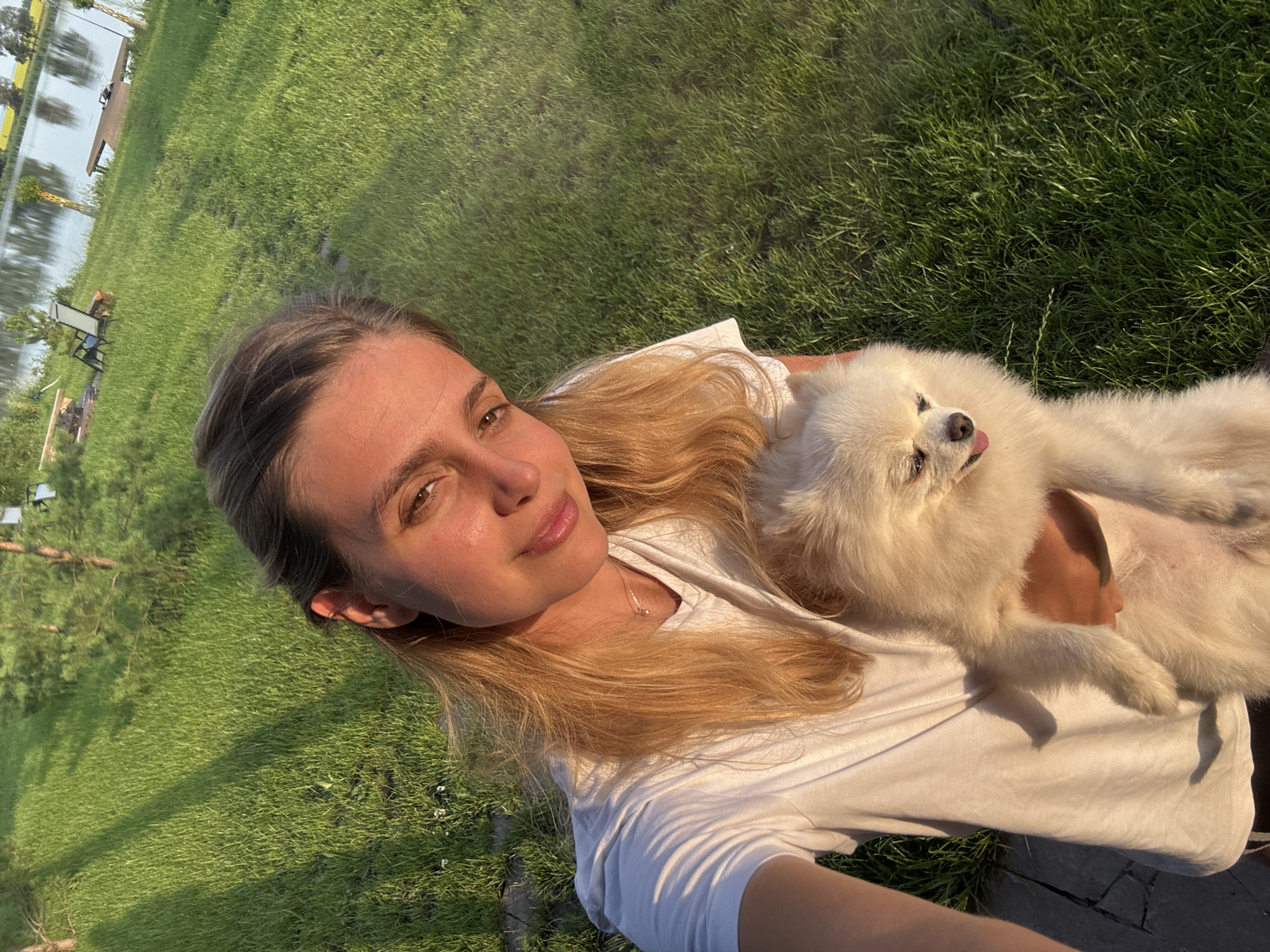 A young woman with long, blonde hair smiles at the camera, holding a fluffy white Pomeranian dog. She is wearing a white t-shirt. The background consists of a lush green lawn, trees, and a body of water, suggesting an outdoor, possibly rural, setting. The sun is casting long shadows, indicating it is either early morning or late afternoon. There are some chairs and a wooden dock visible near the water.