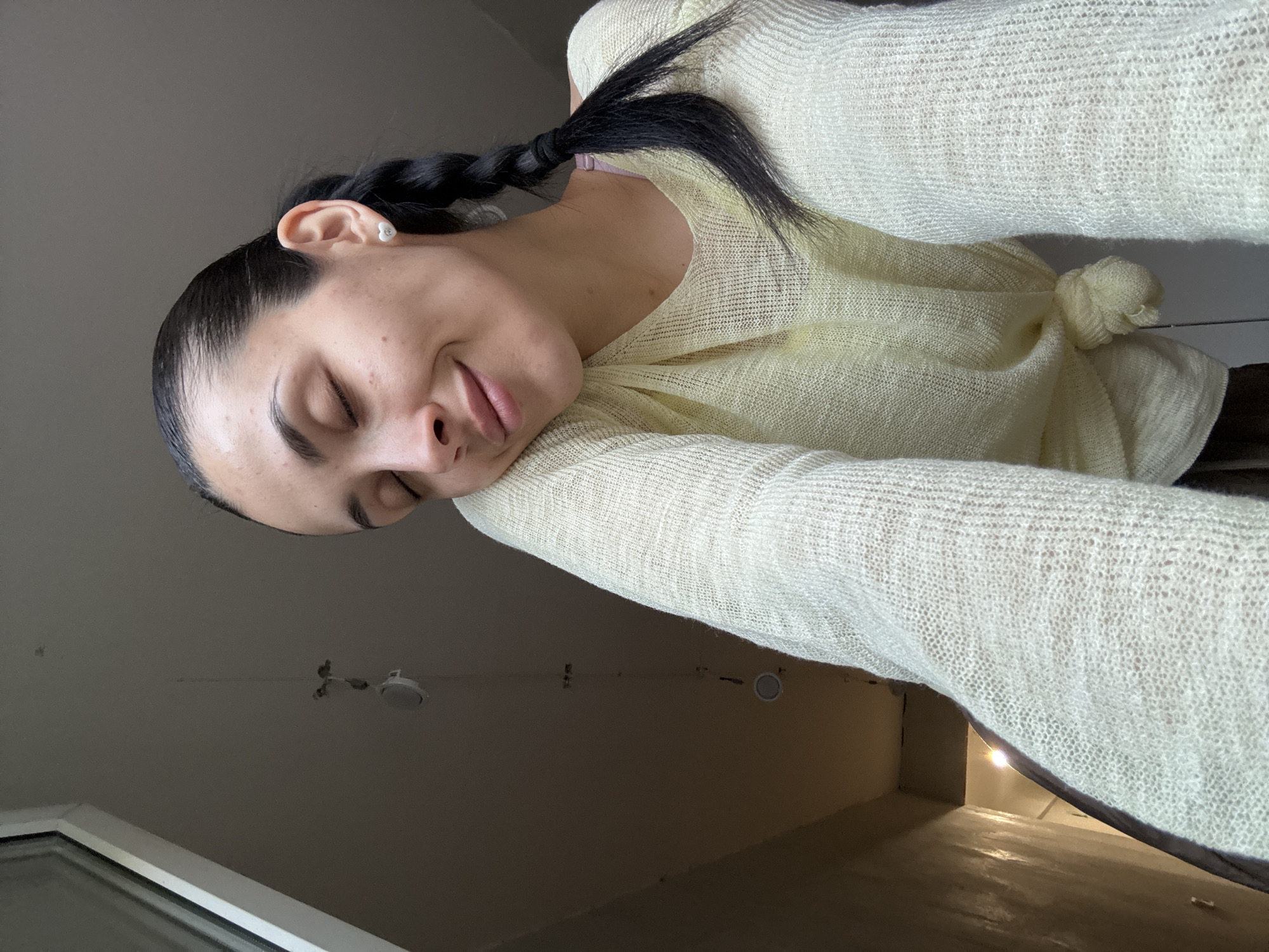 The image features a young adult female captured from a low-angle perspective, looking upwards. She has her eyes closed and displays a gentle smile. Her dark hair is styled into two braids, with one visible resting over her right shoulder, secured by a black hair tie. She wears a small, white, heart-shaped stud earring with a metallic detail in her right earlobe. Her skin shows minor freckles or blemishes.
She is wearing a pale yellow or cream-colored, V-neck, long-sleeved top made of a sheer, loosely knit fabric. The front of the top is tied in a knot at her waist. A hint of a light purple garment is visible underneath her top near her left shoulder.
The background indicates an indoor setting. The ceiling is a light greyish-beige, featuring several white, circular recessed lighting fixtures. A thin cable or wire hangs from the ceiling, connected to a small white, circular object, possibly a speaker or detector. A window frame is partially visible in the upper left corner, and a wall with a slight reflective sheen is seen in the lower left. A brighter light source is visible further back in what appears to be a doorway or hallway in the far lower left.