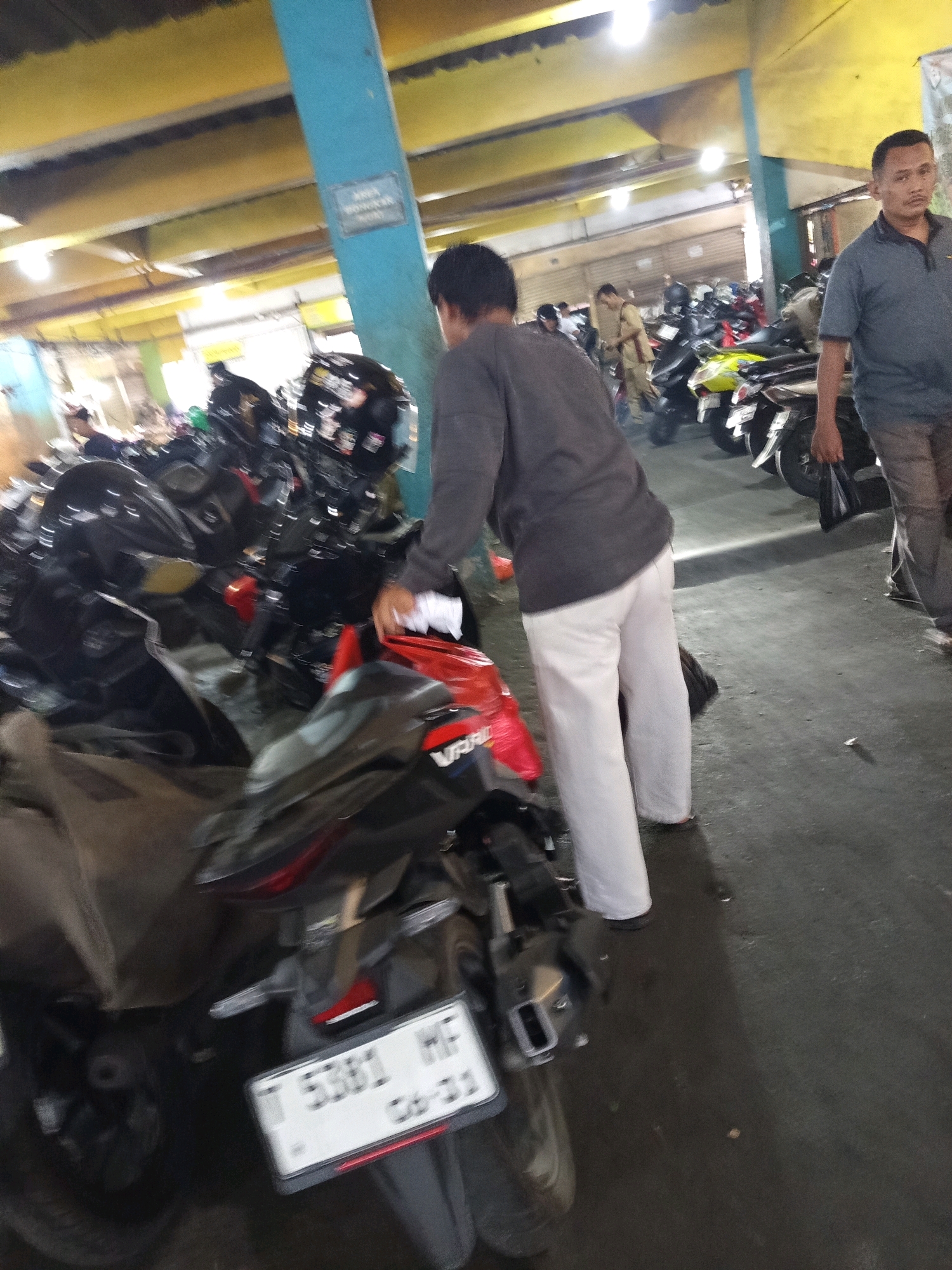 In Karawang, Indonesia, within a dimly lit, multi-story parking facility with a yellowish ceiling and blue pillars, a man in a gray t-shirt and tan pants stands near several parked motorcycles. In the foreground, a person wearing a dark gray long-sleeved shirt and white pants is facing away from the camera, reaching towards the handlebars of a black motorcycle. Another black motorcycle is prominently displayed with a license plate reading "T 5381 WF CB-21". Other motorcycles, including a yellow one, are visible in the background. The floor of the parking facility appears wet in places.
