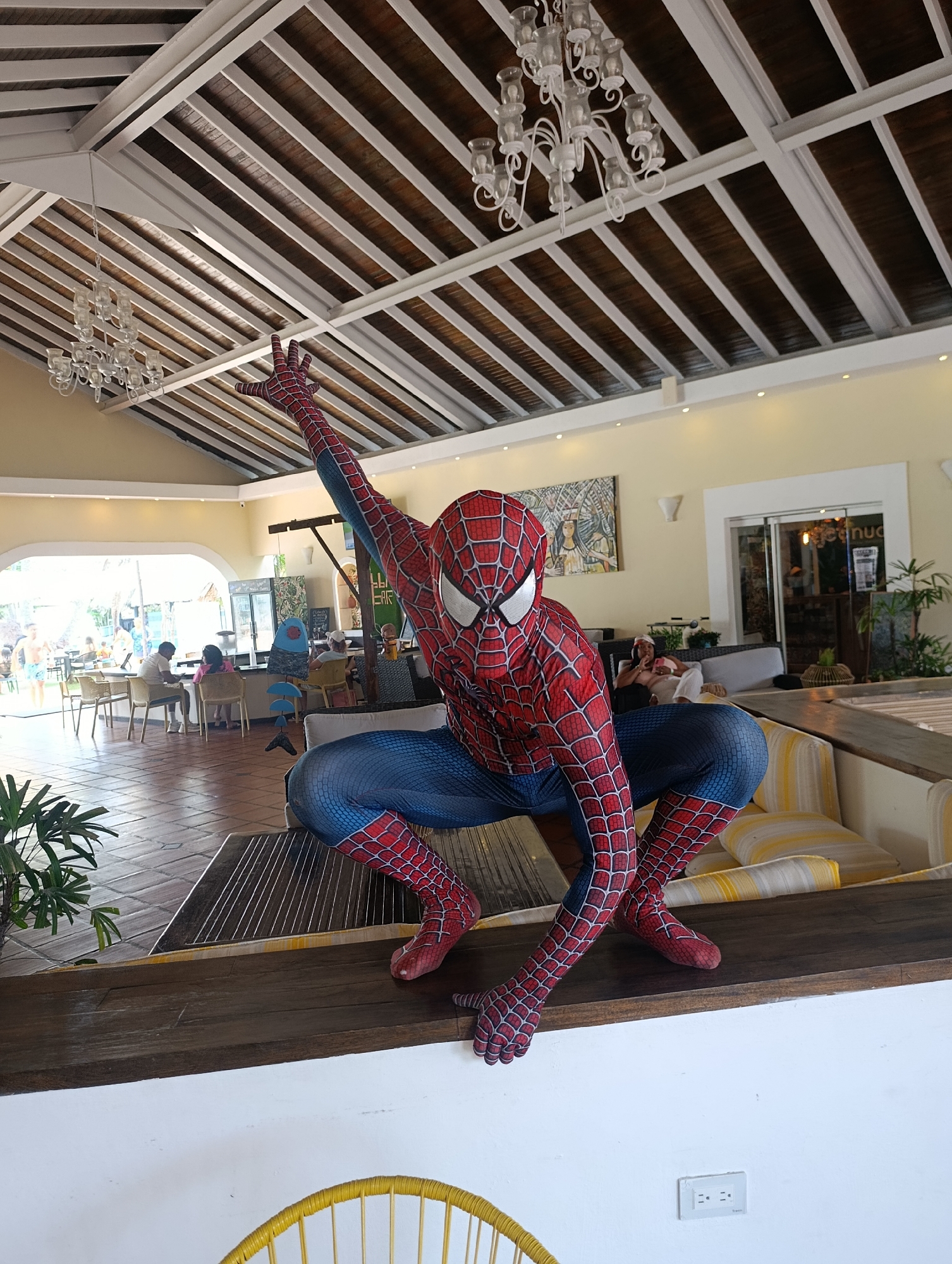 A full-length photograph shows a person dressed in a Spider-Man costume crouching on a dark wooden ledge with one arm raised. The location appears to be the lobby or lounge area of a building. The ceiling is high with exposed beams, and there are two chandeliers. Tables and chairs are visible in the background, along with other people seated. In the very back, a doorway leads to an outdoor area with greenery and people. To the right, a doorway and plants are visible. The wall is light yellow. The base of the ledge is white, with a yellow chair and electrical outlet visible. Without further details, the location cannot be definitively identified in Porlamar, Venezuela.