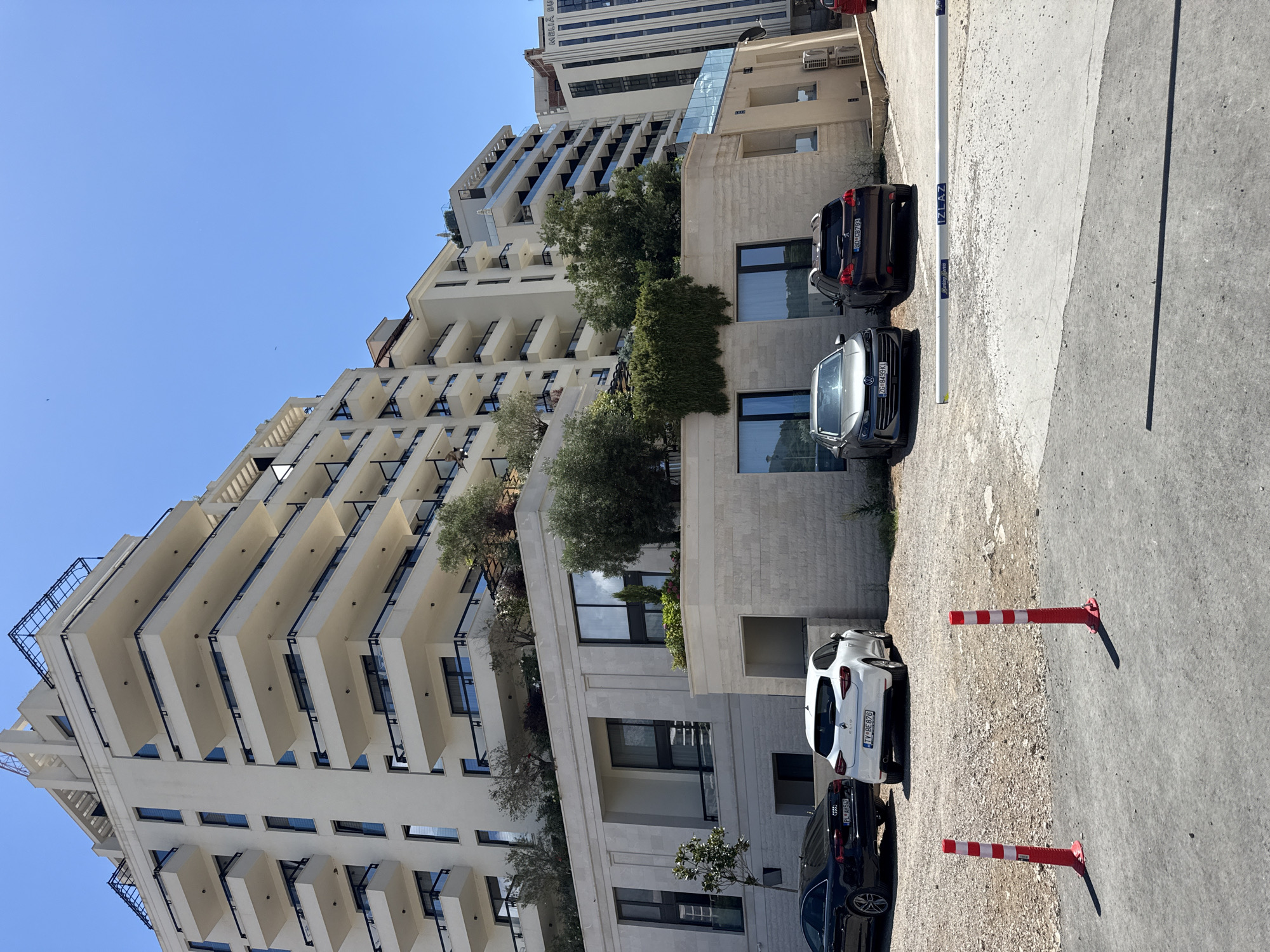 The image shows a modern, multi-story white building with numerous balconies, situated in the city of Rafailovići, Montenegro. In front of the building, there is a parking area with several cars. Specifically, a black Audi sedan is parked on the left, followed by a white Renault Clio and two darker colored SUVs. Red and white striped bollards are placed in the foreground, and a white barrier with "IZLAZ" written on it is visible in the mid-ground, suggesting an exit from the parking area. The building itself features a light-colored stone base with large windows. Greenery, including trees and a vine-covered wall, is integrated into the building's design. The sky is clear and blue. Another tall building with "MELIA DUBROVNIK" written on it is visible in the background to the right.