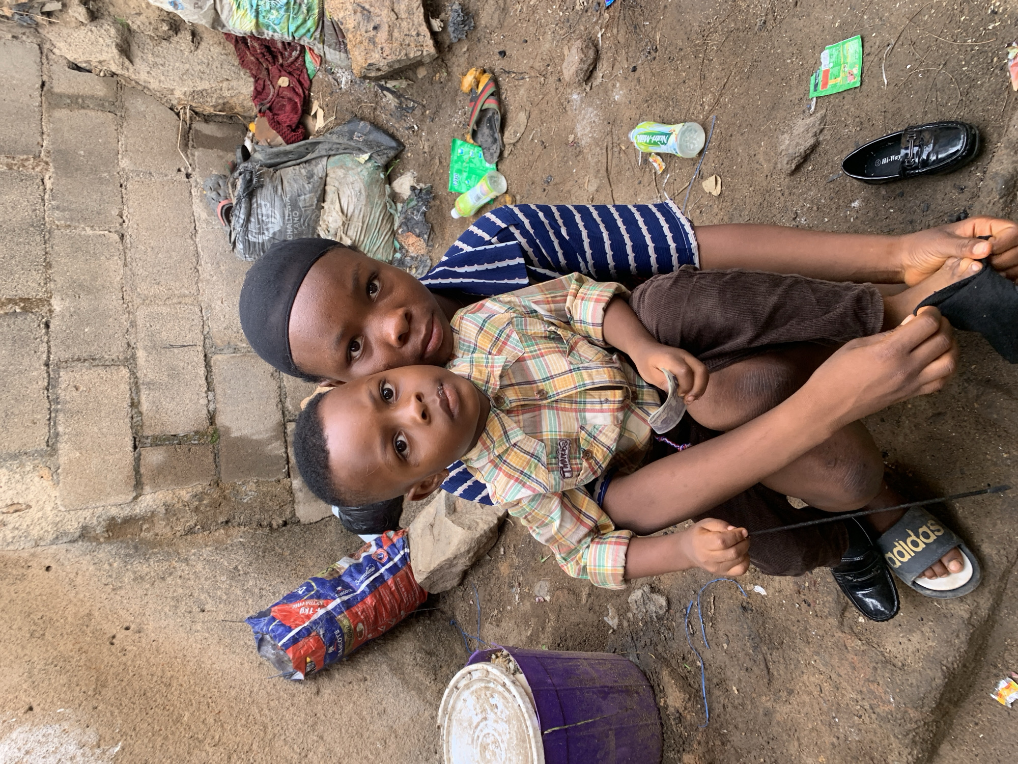 Here is a description of the image:
Two young children are laying on the ground, surrounded by litter and debris. The older child is wearing a black head covering and a blue and white striped shirt. The younger child is wearing a plaid shirt and brown pants. The older child has their arms around the younger child. A bucket and a large bag of Dancote Flour are lying next to the children. A pair of black dress shoes and a pair of Adidas sandals lie near the children's feet. A Hi-Way dress shoe, a bottle of Nutri-Milk, and a green package litter the ground. The location is assumed to be Jos, Nigeria due to the labels of the Dancote bag and the Challenge Kano bag.