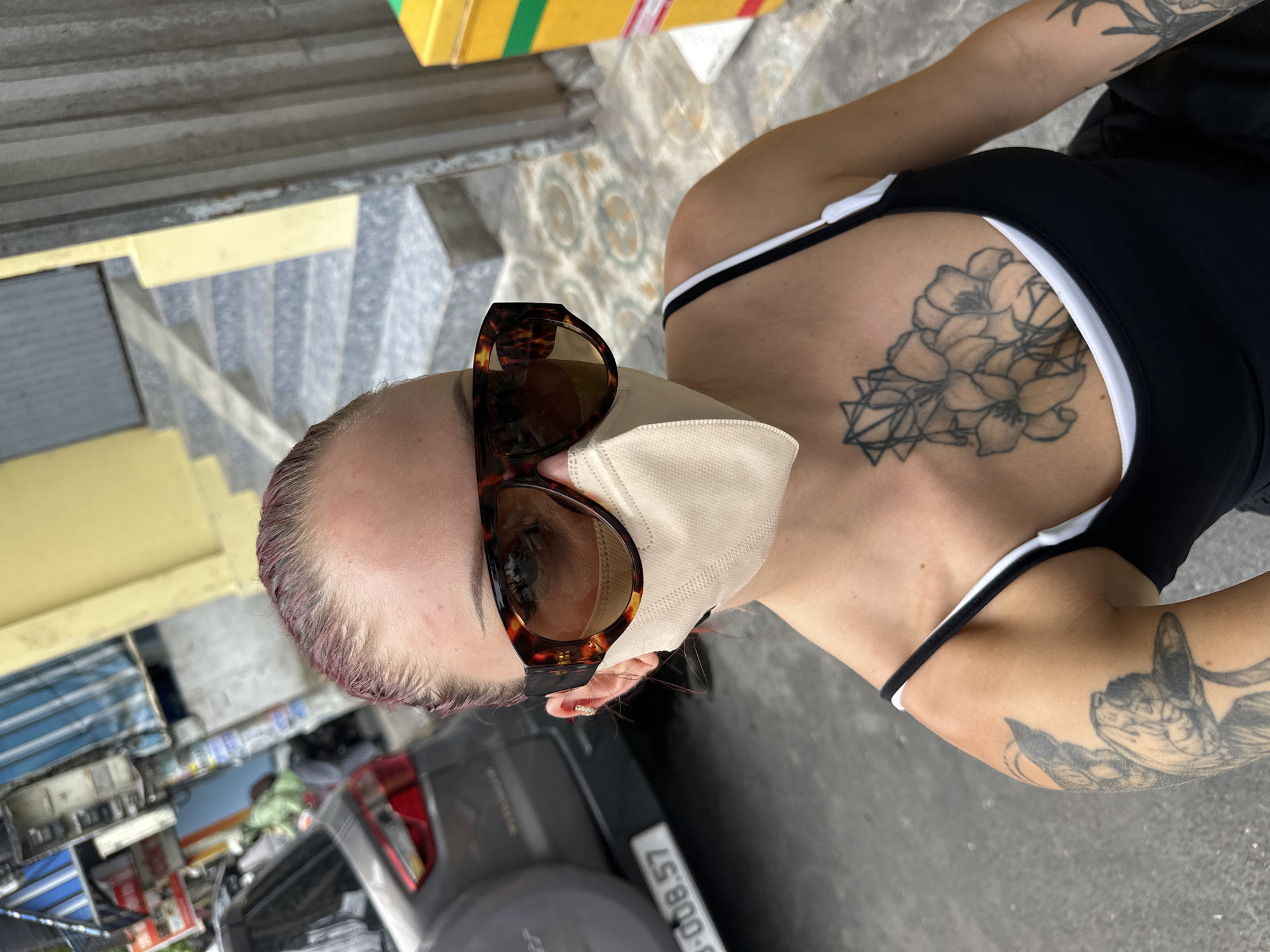 A person wearing large tortoiseshell sunglasses and a light-colored face mask is taking a selfie on a street in Nha Trang, Vietnam. The person has reddish-purple hair and a tattoo of flowers on their chest and an arm tattoo. They are wearing a black tank top with a white undershirt. In the background, a gray car with a license plate that reads "6008.57" is parked on the street, and a yellow and green bin is visible to the right. A yellow building with a set of stairs is behind the person. The street appears to be paved with concrete and decorative tiles.