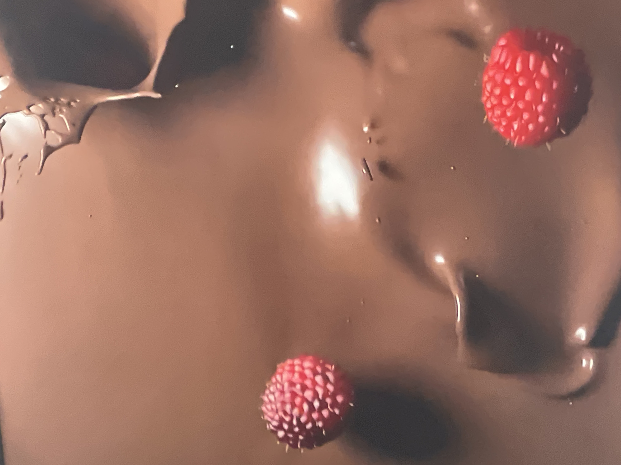 The image shows two bright red raspberries on a glossy brown surface that appears to be melted chocolate. The chocolate has swirls and areas of light reflection, suggesting a liquid or semi-liquid state. The raspberries have a textured surface with small seed-like structures. Location information is not discernible from the image.