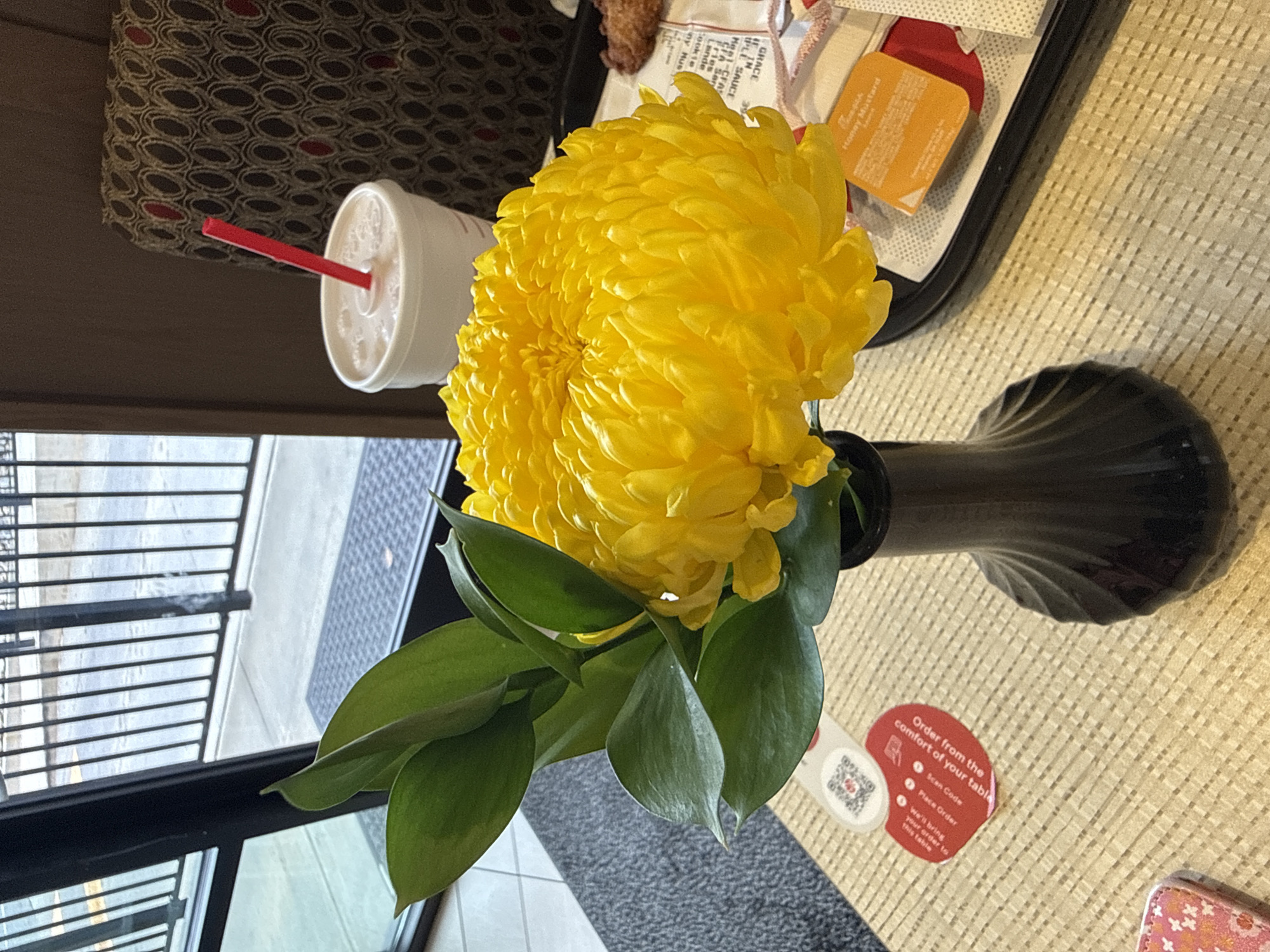 Here is a detailed description of the image:
**Scene:** The image appears to be taken from an overhead perspective on a table within an establishment in Stone Mountain, Georgia, United States. The scene features a table set for dining.
**Objects:**
* A vase containing a large, bright yellow chrysanthemum flower.
* A drink cup with a red straw is visible.
* A Chick-fil-A food tray with takeout items, including a Honey Mustard packet.
* A QR code with the message "Order from the comfort of your table" with the information that you scan the code, place the order, and it will be brought to the table.
* A small patterned purse.
**Environment:**
* The table has a textured surface.
* A window or opening with a view of an exterior sidewalk is visible.
* Seating with patterned upholstery is seen in the background.
**Overall Impression:** The image provides a snapshot of a casual dining experience within an establishment that offers table ordering.