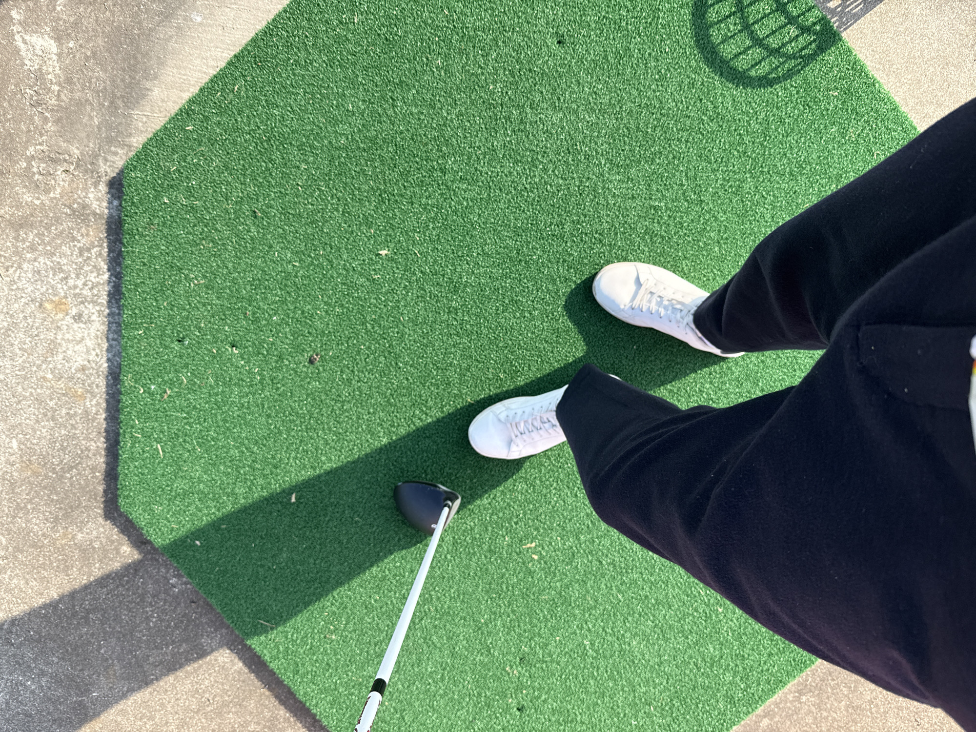A golfer stands on an artificial green golf mat with white shoes and dark pants. A golf club rests on the mat. The mat is octagonal and borders concrete. A shadow pattern is cast in the upper right. The location cannot be determined by visual clues.