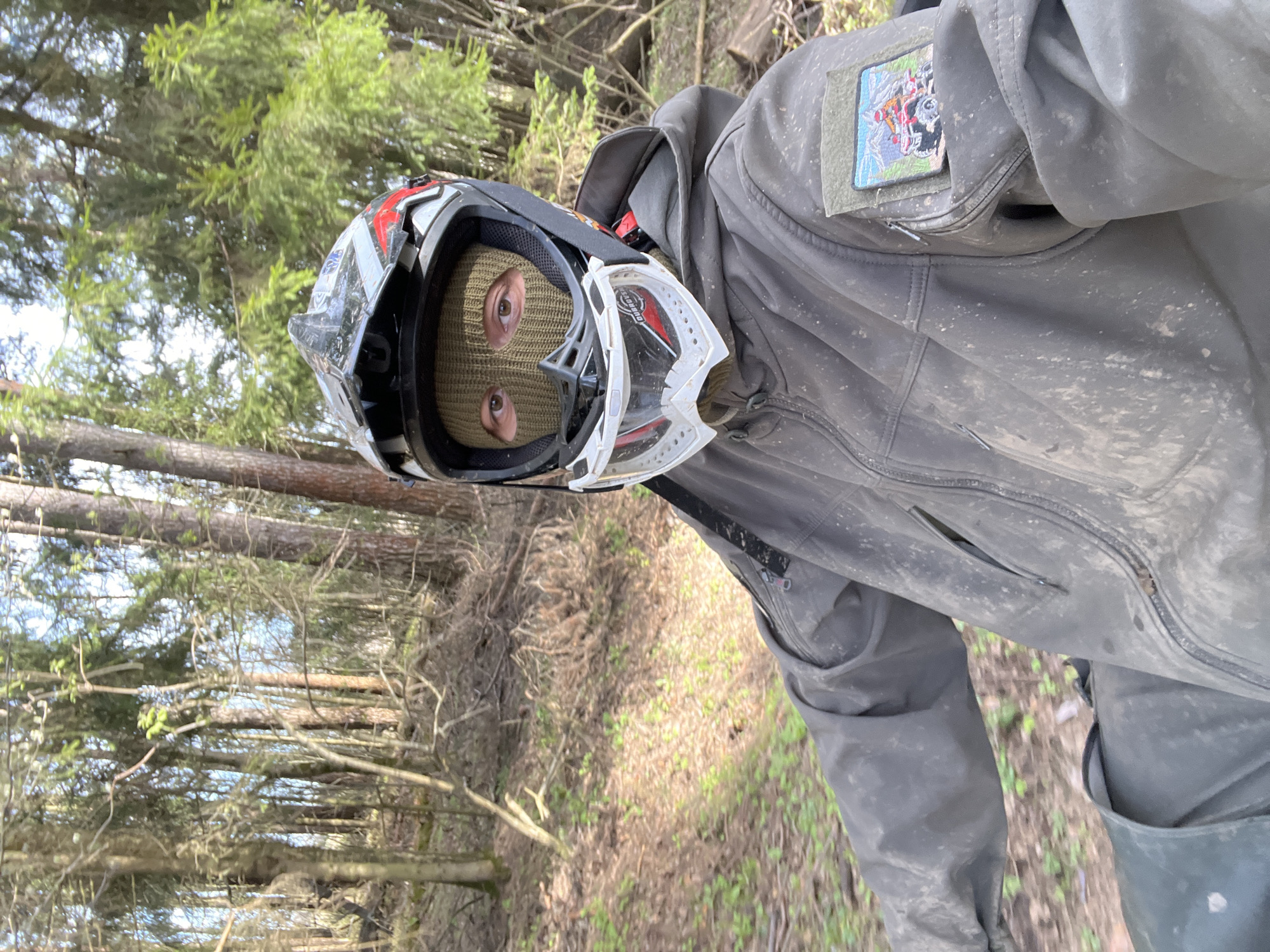 A person wearing a black balaclava, a white and red helmet, and goggles is standing outdoors in a forest environment. They are wearing a dark gray jacket with a patch on the upper left sleeve. The ground is covered with brown leaves and sparse green vegetation, and tall trees are visible in the background. The person is facing the camera, giving the impression of a first-person perspective. The location is likely near Раздолье, Russia, based on provided data.