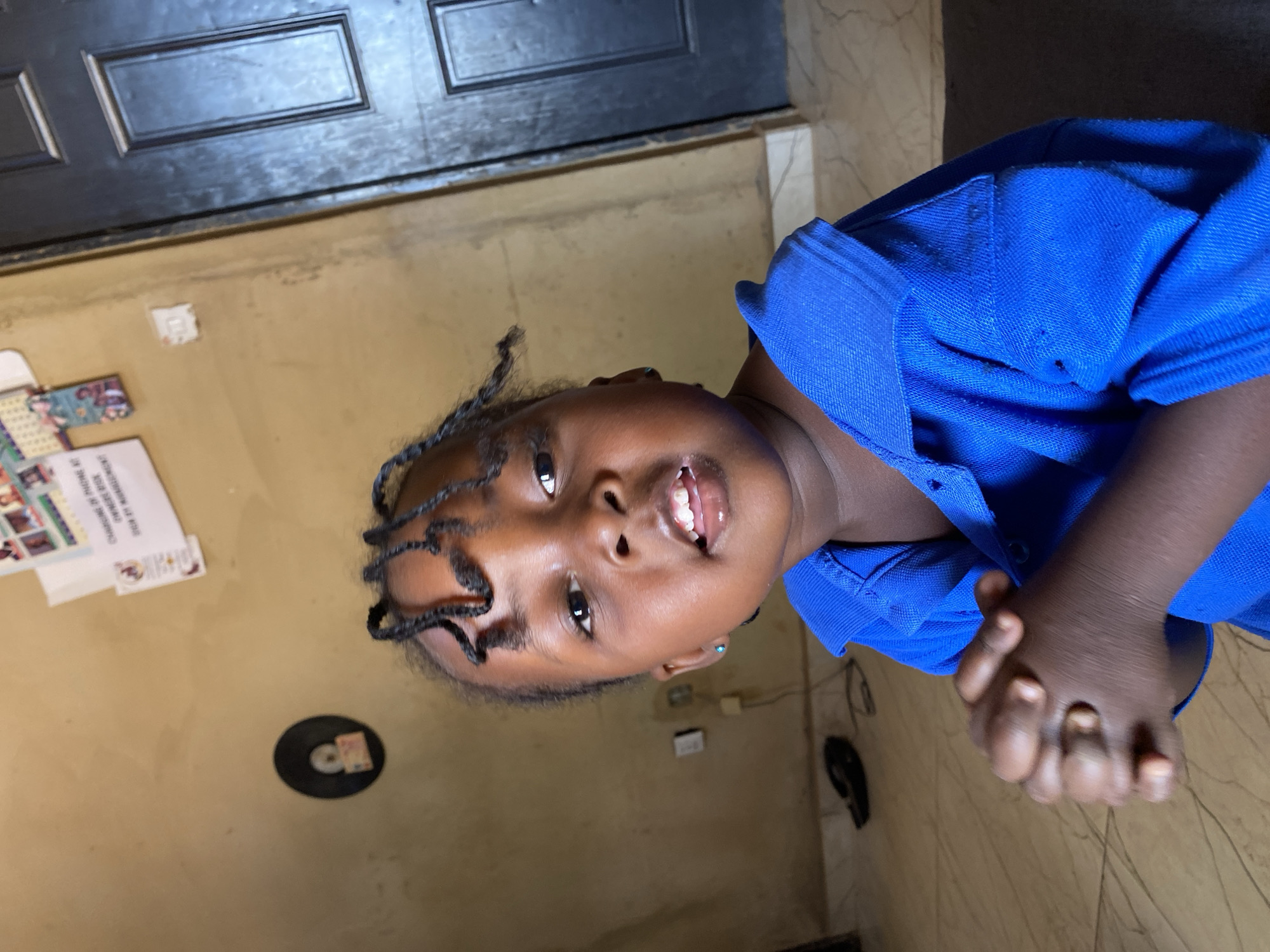 A young child, appearing to be a girl with dark skin and black braided hair, smiles cheerfully directly at the camera, revealing her upper front teeth. She wears a bright royal blue polo shirt with a collar and buttons, and a small blue stud earring is visible in her left ear. Her hands are clasped together in front of her.
The setting is indoors, likely within a home in Kuru Dungus, Nigeria. The background features light beige or tan walls and a patterned floor, possibly tiled. Behind and above the child, a dark brown or black wooden door with rectangular panels is visible, suggesting a doorway. On the wall to the left of the child, several papers or cards are affixed, with partially legible text that includes phrases like "HOME OF" and "CHANGE OF FUTURE". A small black circular object, possibly a CD or vinyl record, rests on the floor to the child's left, with a small white item on top of it. An electrical outlet or switch is also discernible on the wall further back. The child's expression conveys happiness and friendliness, captured from a slightly low camera angle.