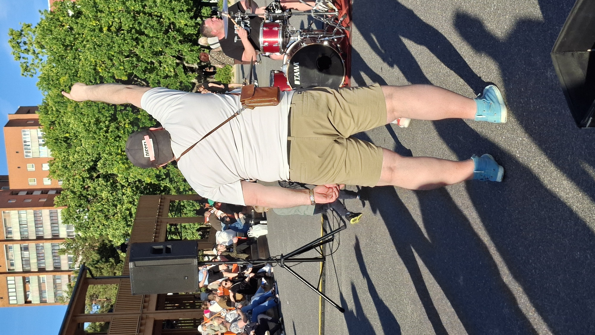 The image shows an outdoor scene with several people and a drum set. A man in the foreground, seen from the back, is wearing a white t-shirt, khaki shorts, a baseball cap, and light blue sneakers. He is pointing his right arm upwards and outwards. A small brown pouch is attached to his belt. Behind him, a drummer is seated at a red drum set. The setting appears to be a public space, possibly an outdoor event or concert, with a crowd of people seated in the background. There are trees and a multi-story brick building visible in the background. The lighting suggests it is daytime, likely sunny given the strong shadows cast on the ground. The overall atmosphere seems casual and energetic, possibly during a performance. The word "DIESEL" is visible on the baseball cap worn by the man in the foreground.