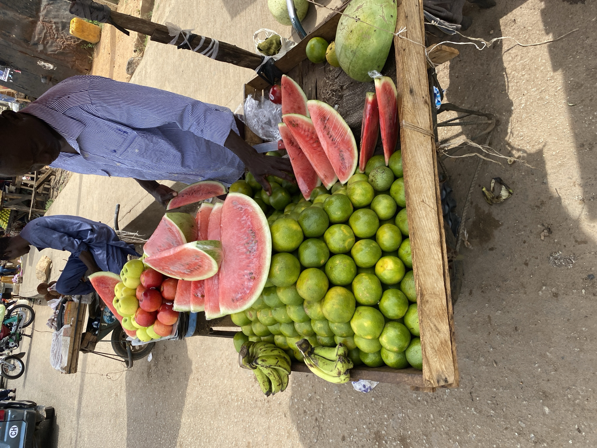 The image provides an overhead view of an outdoor fruit stall or market stand in what is described as Bukuru, Nigeria. A vendor, wearing a blue and white striped long-sleeved shirt, stands behind a wooden cart or tricycle-mounted display laden with various fresh fruits. His dark-skinned hand is visible near the fruit, suggesting interaction or arrangement.
The main display features a prominent arrangement of freshly sliced watermelons, some pieces wrapped in clear plastic. A large section of the cart is dedicated to a substantial pile of small, green citrus fruits, likely unripe oranges or limes, stacked in organized rows. Below these, several bunches of green and yellow bananas are visible. Another part of the display holds a collection of red and green apples. A few larger, whole green fruits, possibly watermelons or gourds, are also visible towards the back of the stall.
Another individual, dressed in a blue garment, is partially visible to the left, near a smaller trolley displaying more fruit. The setting appears to be a street or market area with concrete and dirt ground. In the background, rudimentary structures and parts of motorcycles or scooters are visible. A sticker or license plate bearing the letters "GH" is present on a dark vehicle in the lower-left corner. The bright sunlight indicates it is daytime, casting clear shadows. The scene depicts a typical, active market environment.