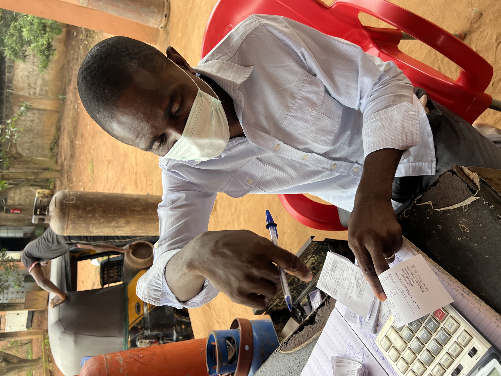 A Black man, wearing a light blue pinstripe button-down shirt and a white face mask, is seated in a red plastic chair at a dark, weathered table outdoors in Nkpor, Nigeria. He is holding a blue and black pen and pointing at a white receipt with his right index finger. The receipt, which appears to be a fuel station receipt, is positioned over a stack of other papers and a calculator. Another receipt, also white, with "TRIGAS" and "RECEIPT INVOICE" printed on it, is held between his thumb and forefinger. The calculator has silver and grey keys. In the background, another Black man, wearing a black t-shirt and grey shorts, is leaning into a yellow and grey auto-rickshaw, holding a gas cylinder. There are several large, orange and grey gas cylinders visible in the foreground and background, as well as stacks of smaller blue gas canisters. The ground is made of reddish-brown earth. The scene appears to be a retail setting, possibly a gas station or a fuel vendor.