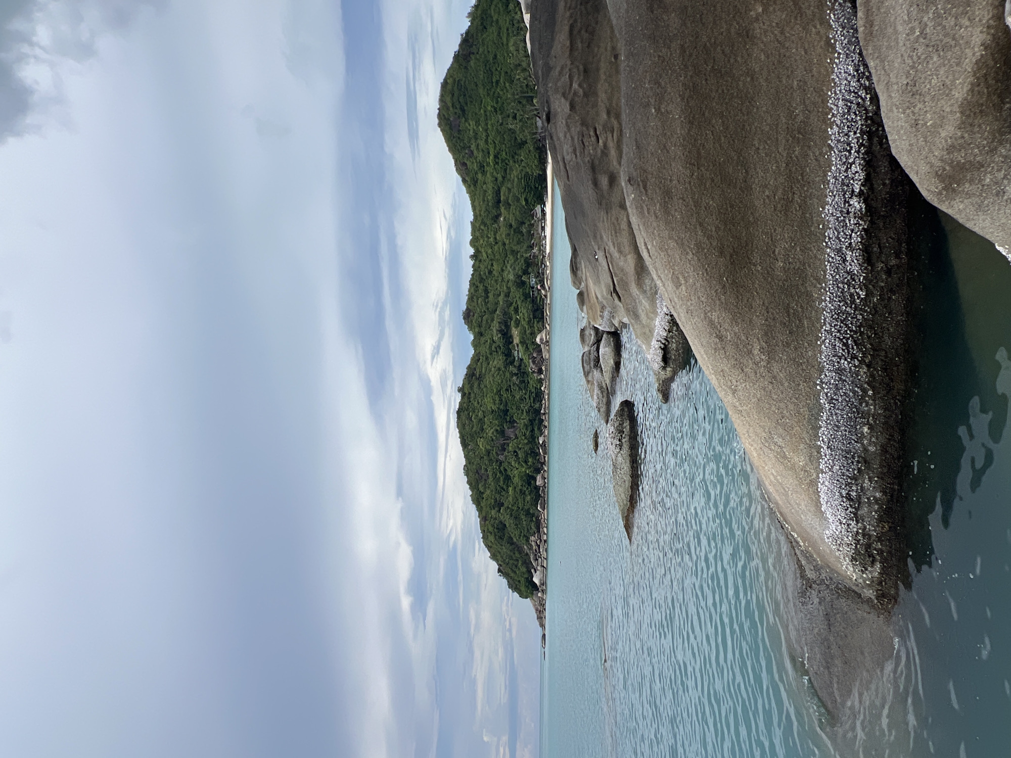 The image depicts a coastal scene at Baan Chaweng Noi, Thailand. In the foreground, large, weathered boulders are partially submerged in calm, turquoise water. These rocks are encrusted with small, white barnacles, particularly along the waterline. The water exhibits gentle ripples. In the middle ground, a stretch of sandy beach is visible, backed by a densely forested, green hillside that slopes upward to form a prominent headland. The sky above is overcast with layers of grey and white clouds, suggesting a diffused light. No people or specific actions are identifiable.