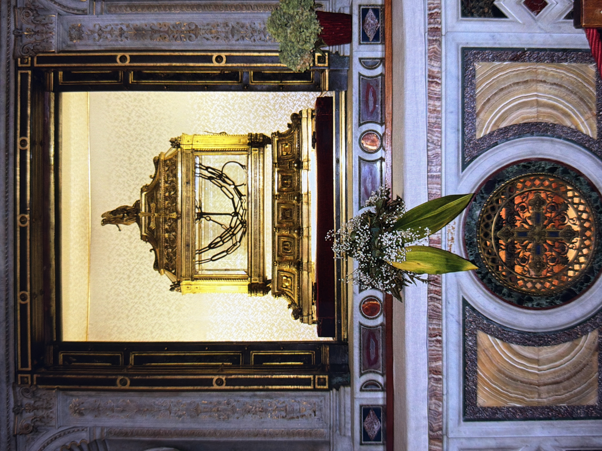 The image depicts an ornate, gilded reliquary or monstrance, likely an artifact of religious significance, set within an elaborate architectural interior. The object is encased in glass and surrounded by decorative gold metalwork, featuring intricate scrollwork and architectural elements. Inside the reliquary, a dark, cross-shaped object is visible. The reliquary is mounted on a richly decorated surface, possibly a wall or altar, adorned with various marbles and intricate carvings. To the right of the reliquary, a circular decorative element with a metal grille and a cross motif is set into the marble. A bouquet of small white flowers and green leaves is placed to the left of this circular element, partially obscuring the reliquary. The overall scene suggests a sacred space, possibly a church or cathedral, given the opulent decoration and the nature of the central object. The lighting appears to be artificial and warm, illuminating the golden hues of the reliquary and the surrounding decor. The perspective is from above, looking down at the object and its surroundings. No people are visible, and no text is discernible in the image. The context of Kyiv, Ukraine, implies a historical or cultural setting within a Ukrainian religious institution.