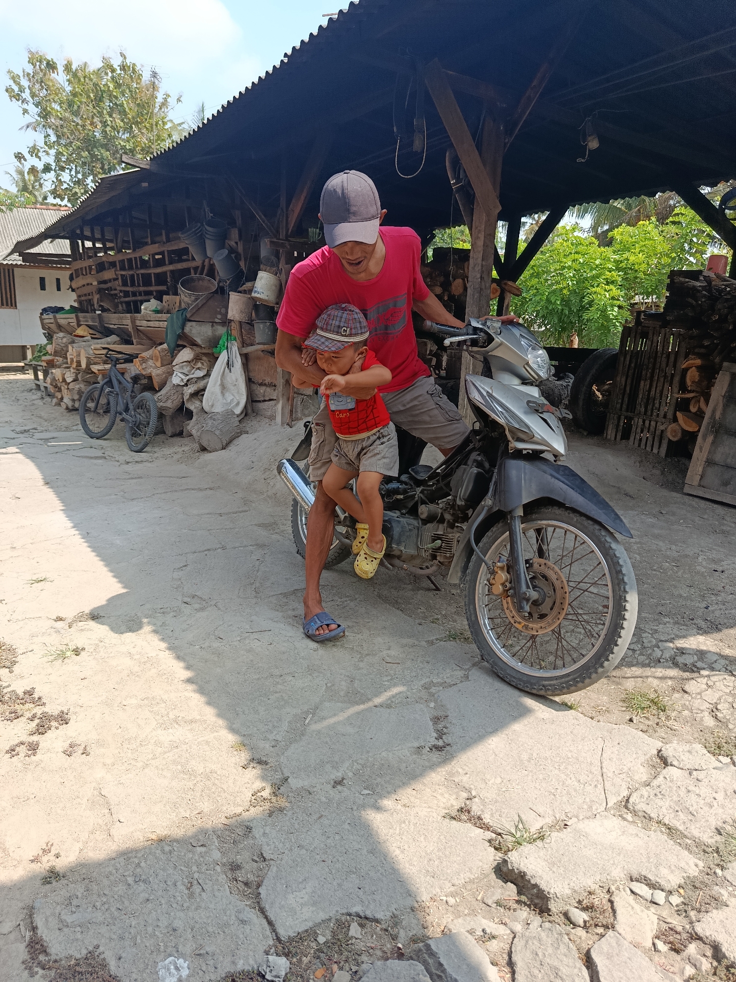 In Sarwadadi, Indonesia, a man in a red t-shirt and a grey cap holds a small child wearing a red shirt and a checkered cap. The man is positioned next to a silver and black motorcycle, with his right hand on the child's back and his left hand near the motorcycle's handlebars. The child is seated on the motorcycle, facing the man. Both are outdoors on a dirt and concrete ground, with shadows cast by overhead structures. In the background, there is a wooden structure with a corrugated metal roof, piled with logs and various objects, including buckets and bags. A bicycle is parked to the left of the motorcycle. Lush green trees and foliage are visible further in the background. The scene appears to be a rural or semi-rural setting, possibly a workshop or storage area.