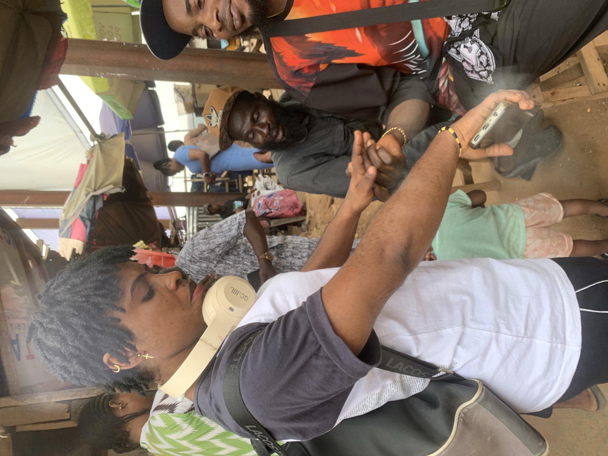 In the city of Shagamu, Nigeria, at an outdoor market, a young woman with dreadlocks and headphones around her neck is the central figure. She wears a black and white t-shirt and a backpack. To her right, a man with a full beard and a brown hat sits with his mouth open, seemingly mid-conversation. He is wearing a dark shirt and bracelets. Beside him, another man wearing a colorful orange, black, and red shirt and a baseball cap is looking towards the camera. A young child in a light green t-shirt and patterned shorts is visible in the background, as are other market-goers and stalls with umbrellas. The ground is dirt and there are wooden structures and chairs. The scene captures a moment of interaction and daily life within the bustling market.