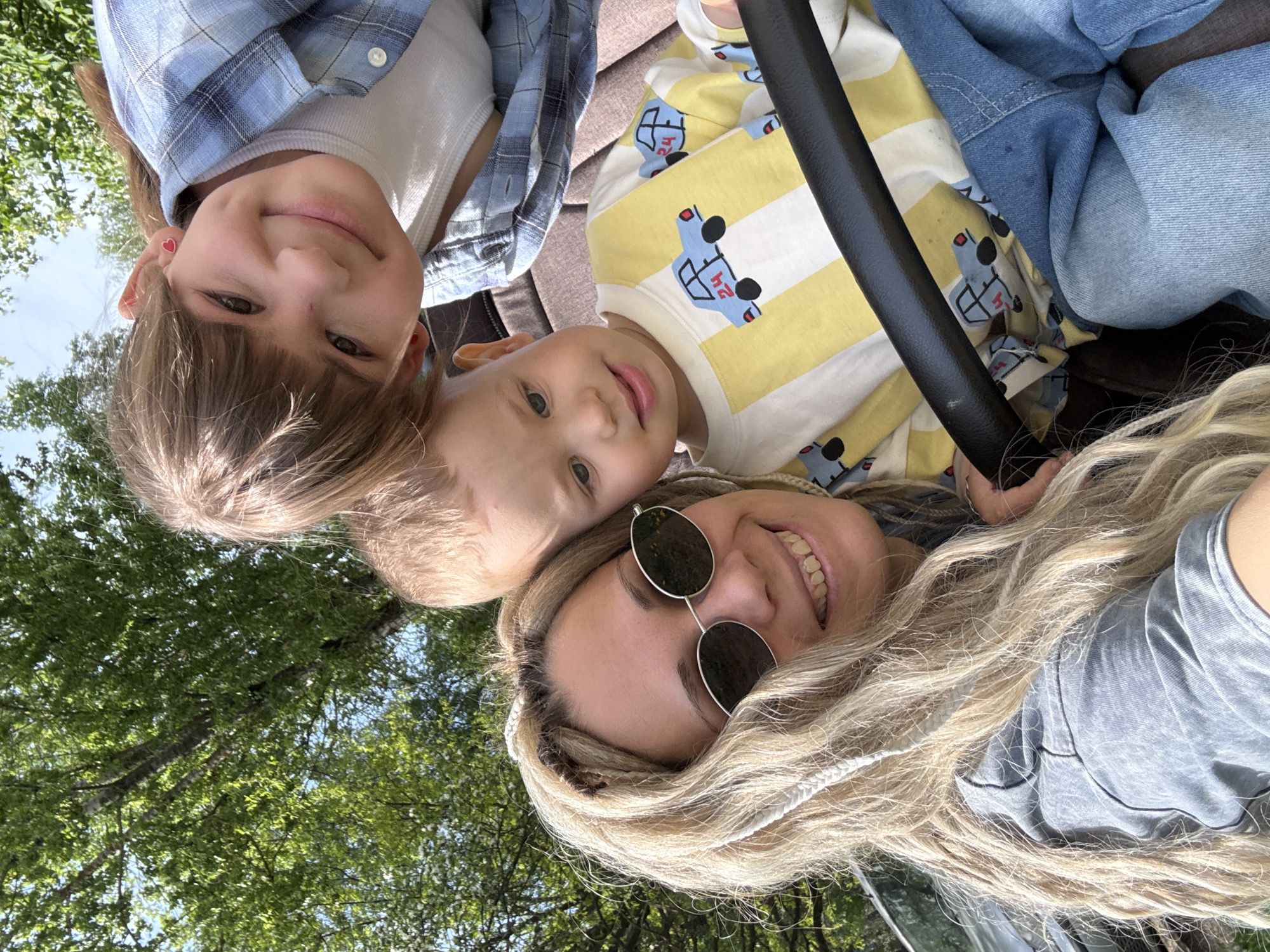 A low-angle selfie shows a woman with blonde braided hair, wearing round sunglasses and a grey t-shirt, with two children. The child on the left has brown hair, is wearing a white tank top and a blue plaid shirt. The other child, in the middle, has blonde hair and is wearing a yellow and white striped shirt with a car pattern. The people are laying down or looking directly at the camera. In the background there is green foliage. There is a black bar running in front of the woman's face. The location is possibly Rusanovo, Russia.