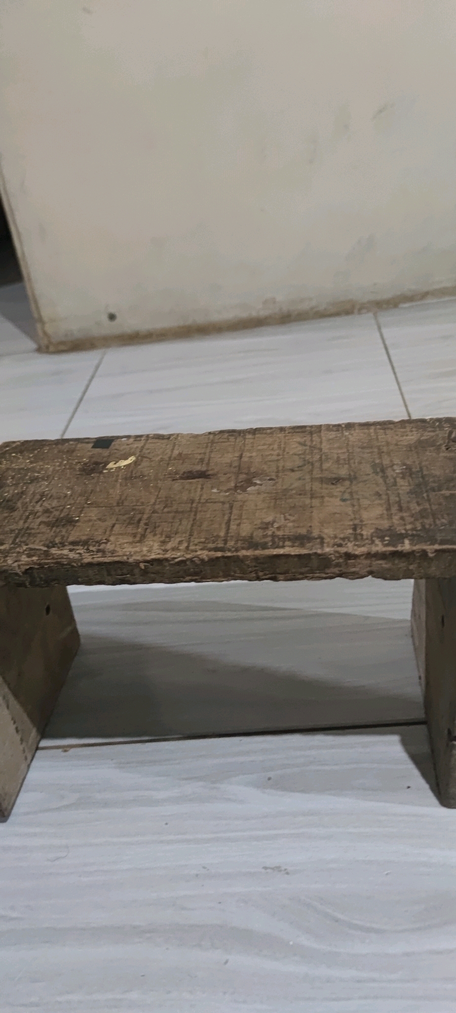 A low-angle shot captures a rustic, wooden stool placed on a tiled floor in Gambarsari, Indonesia. The stool, with its weathered, rough-hewn surface and simple block legs, appears to be handmade. A pale wall forms the background, with a slight shadow cast by the stool onto the floor. The lighting is dim, suggesting an indoor environment. There are no people or discernible actions taking place in the scene.
