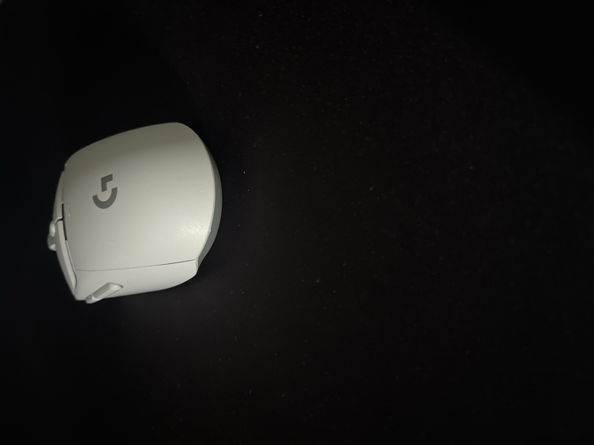 The image features a white Logitech gaming mouse with a grey logo on a dark, textured surface, likely a mousepad. The mouse is positioned on the left side of the frame, angled slightly. Its smooth, ergonomic design is visible, including buttons and a scroll wheel. The background is predominantly black, with subtle speckles suggesting dust or texture on the surface. The lighting is dim, casting a soft glow on the mouse, and it appears to be an indoor setting, likely a desk area. There are no people or specific location cues besides the general context of Varna, Bulgaria. The image focuses solely on the object itself, without any discernible activity or event taking place. The Logitech logo is the only visible text.