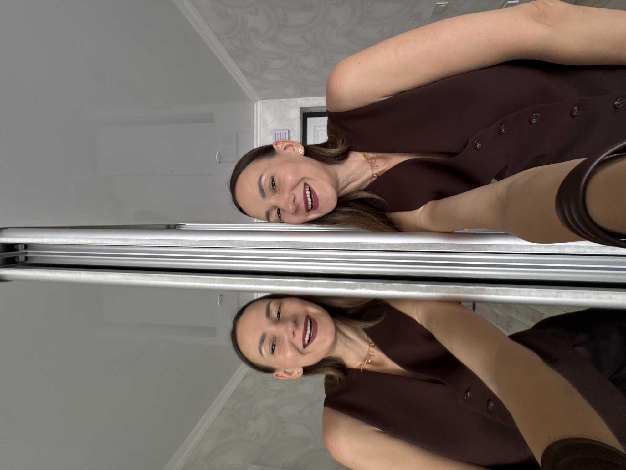 This media file displays a cheerful woman captured in a selfie, presented with a 90-degree clockwise rotation. The image features the woman smiling broadly, revealing white teeth, and wearing dark red lipstick. She has long, dark brown hair and is adorned in a dark brown, sleeveless button-down top or vest. A prominent gold-colored necklace with individual letter charms spelling out "L O V E" (and possibly more, partially obscured) hangs around her neck, complemented by a small gold earring in her right ear.
Below her, a highly reflective surface, likely a large mirror with a silver-toned frame, captures a clear, inverted reflection of the woman and her immediate surroundings. The background behind the woman reveals an indoor setting with a light-colored wall featuring a subtle, patterned wallpaper. A white door frame is partially visible, along with a small white rectangular fixture on the wall, possibly a thermostat or switch. The lighting suggests it is daytime. The woman's expression conveys happiness and contentment. This scene is set indoors, possibly within a home or hotel in Sochi, Russia.