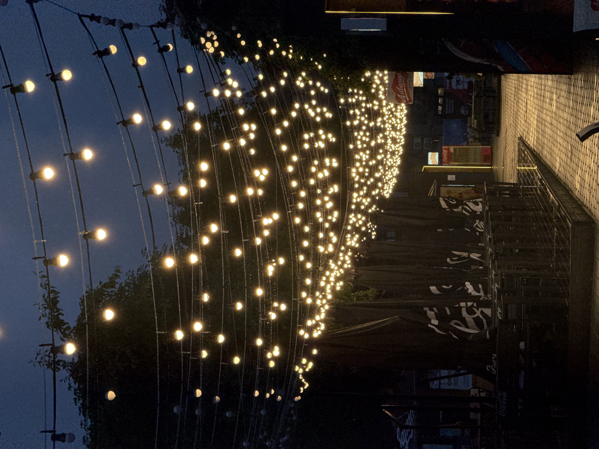 The image depicts an outdoor seating area at night, illuminated by numerous strings of warm-toned fairy lights. The lights are draped across multiple wires, creating a canopy effect overhead. In the foreground, dark fabric-covered umbrellas are neatly arranged, possibly indicating a restaurant or cafe. The ground is paved with light-colored tiles, reflecting the overhead illumination. In the background, there are glimpses of buildings and signs, some of which appear to be in Cyrillic script, suggesting an Eastern European location. One sign clearly reads "Coca-Cola." The overall atmosphere is one of a cozy, illuminated public space in the evening. The city of Minsk, Belarus, is a possible location given the Cyrillic script and the general urban setting.