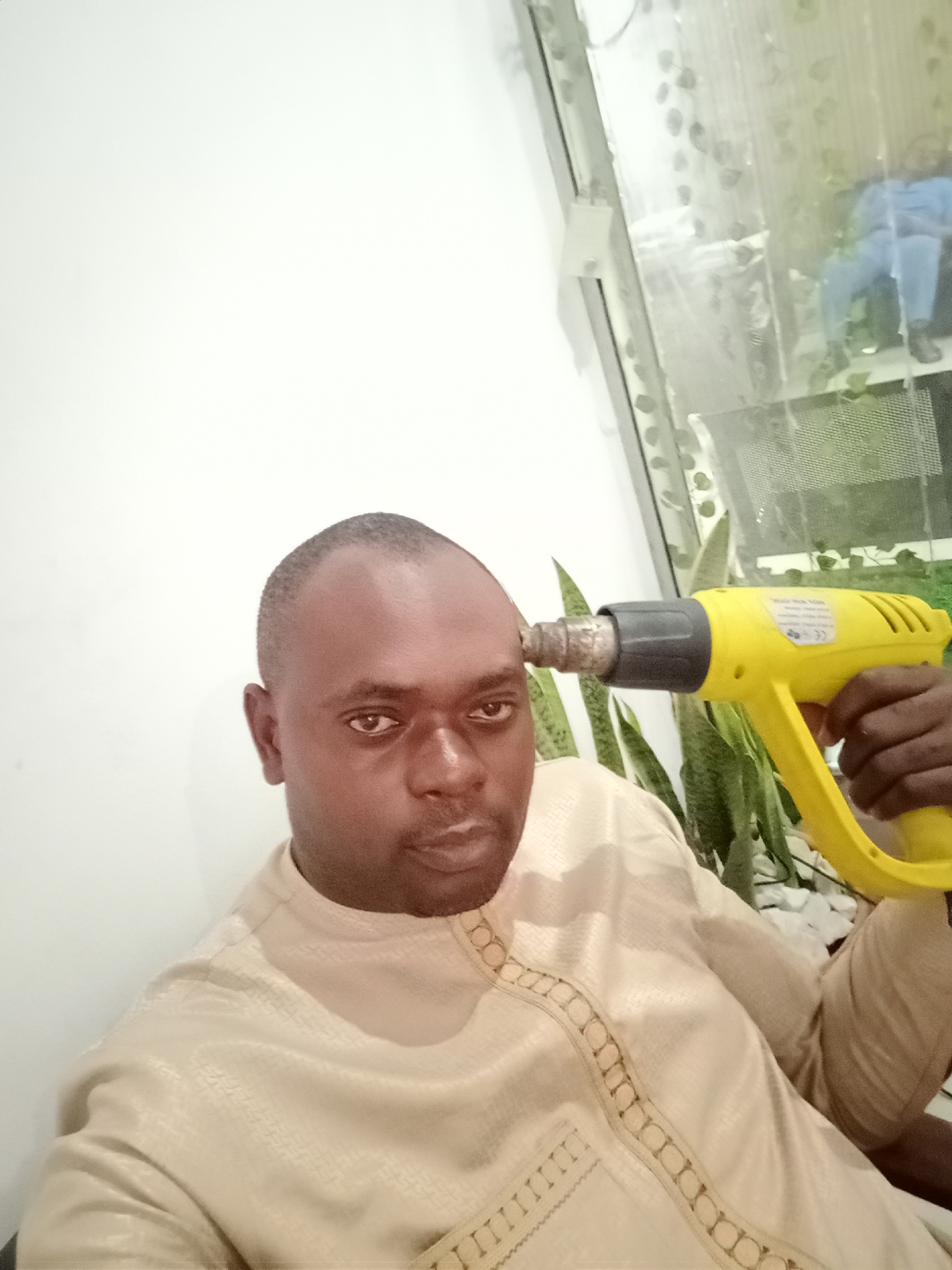 A dark-skinned man holds a yellow heat gun to his head in an indoor setting. The man is wearing a light beige shirt with decorative embroidery along the neckline. Behind him, there's a window showing greenery and a seated person. The location is in Gwarinpa, Nigeria.