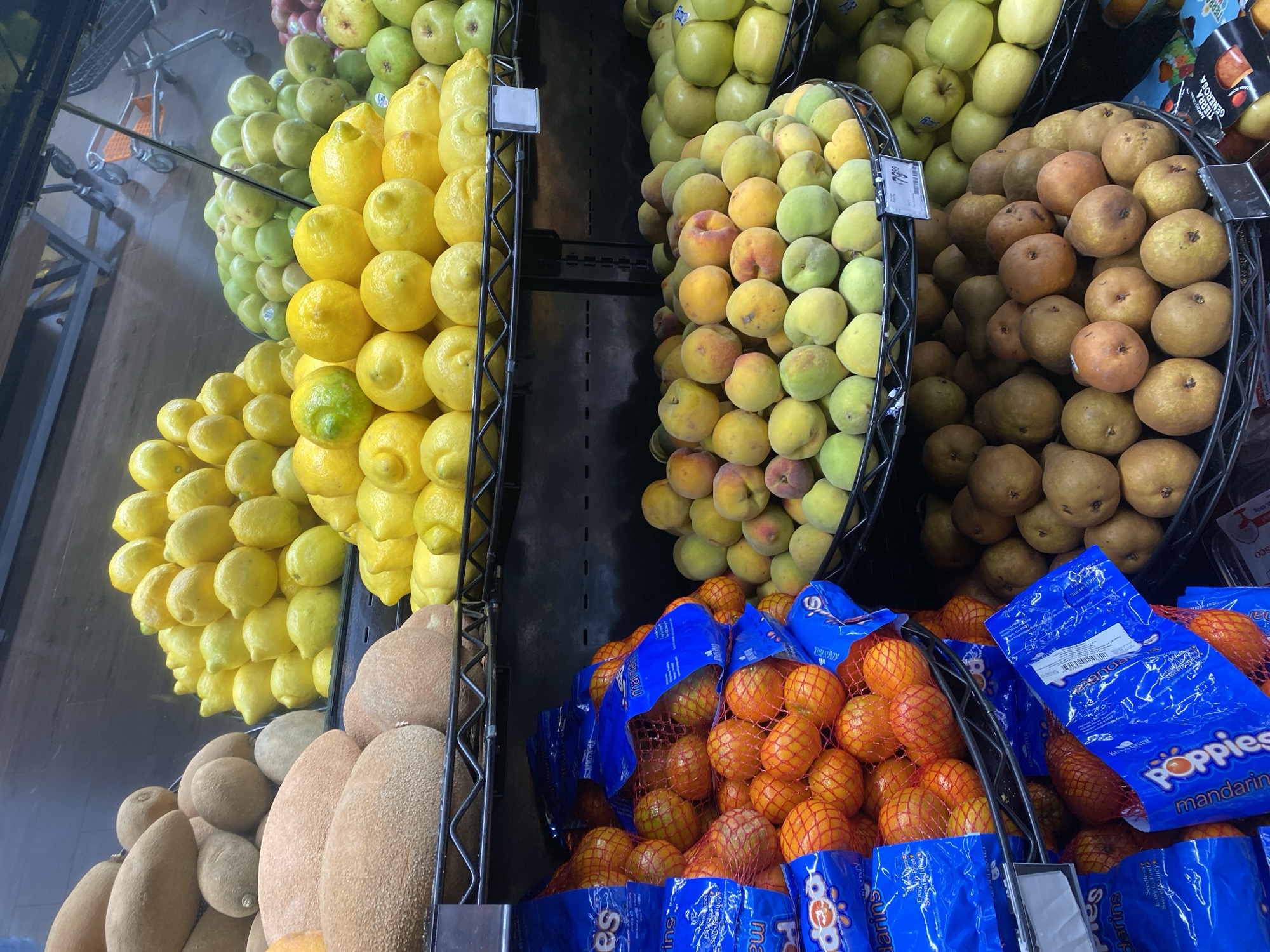 The image shows a produce display at a grocery store. From left to right, there are bins or shelves filled with green apples, lemons, chico fruits, yellow peaches, pears, and bags of tangerines. The "Poppies" brand of mandarin oranges is visible. A small shopping cart can be partially seen in the background. The scene seems to be indoors, likely in the produce section of the store. It's impossible to establish the location of the city of San Bernardino Tlaxcalancingo, Mexico.