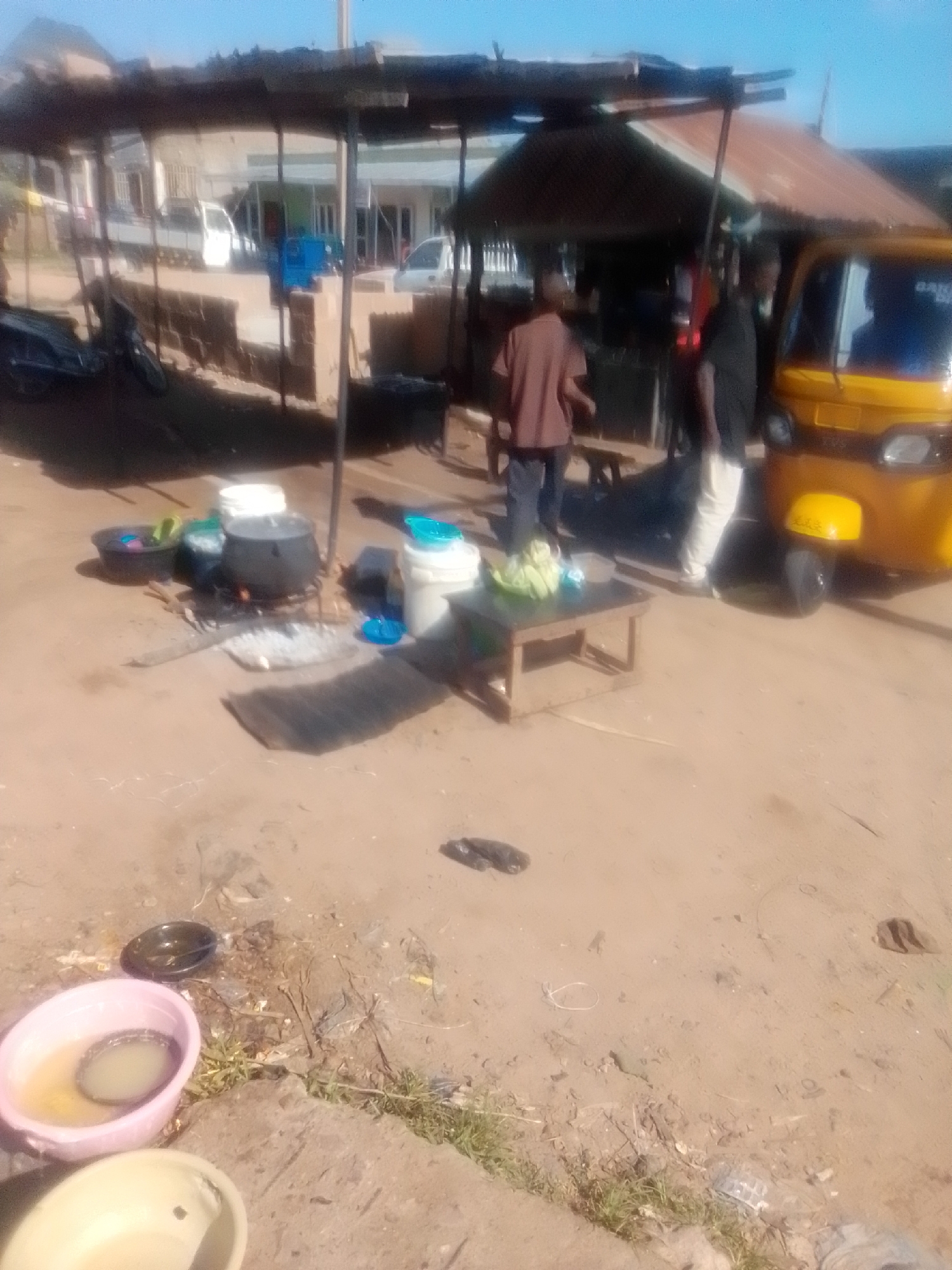 This image captures a bustling scene in Pantame, Nigeria, likely a roadside market or food stall. In the foreground, several large plastic basins are scattered on the dusty ground, one containing murky water and another empty. Further back, a fire is burning under a large pot, with other buckets and containers around it, suggesting food preparation is underway. Two individuals are visible in the mid-ground, one wearing a brown shirt and the other a black shirt, appearing to interact with each other or manage the stall. A bright yellow three-wheeled auto-rickshaw is parked nearby, adding to the commercial atmosphere. The background reveals more stalls and buildings, hinting at a densely populated urban or semi-urban environment. The overall impression is one of daily life and commerce in a Nigerian city.