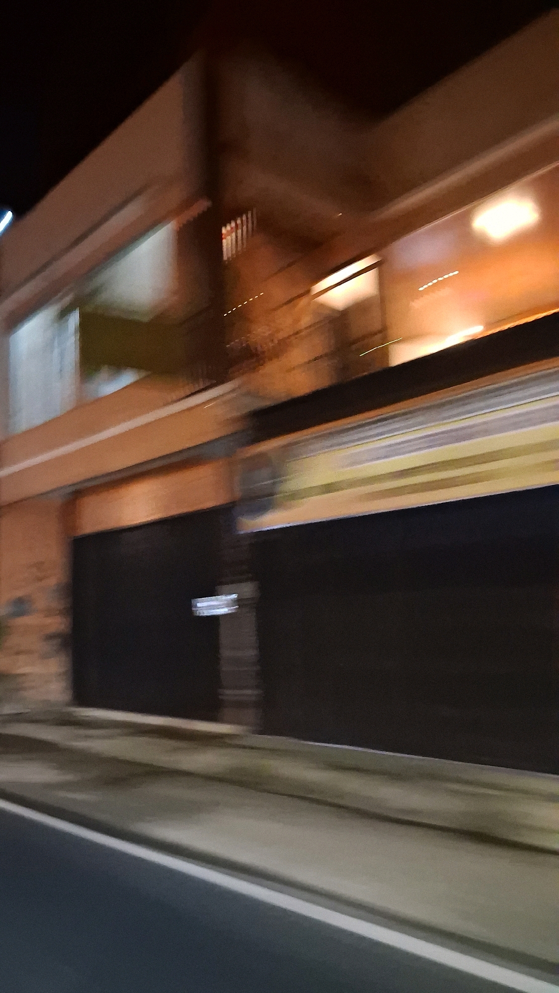 The image depicts a blurry, motion-distorted view of a building at night, likely taken from a moving vehicle on a street in Berbah, Indonesia. The building appears to be a commercial establishment with a dark facade and illuminated windows on an upper level. The lighting suggests artificial light sources, possibly interior lights and streetlights. The ground in front of the building is a sidewalk or paved area, and a road is visible in the foreground. The overall impression is one of urban activity captured during nighttime.