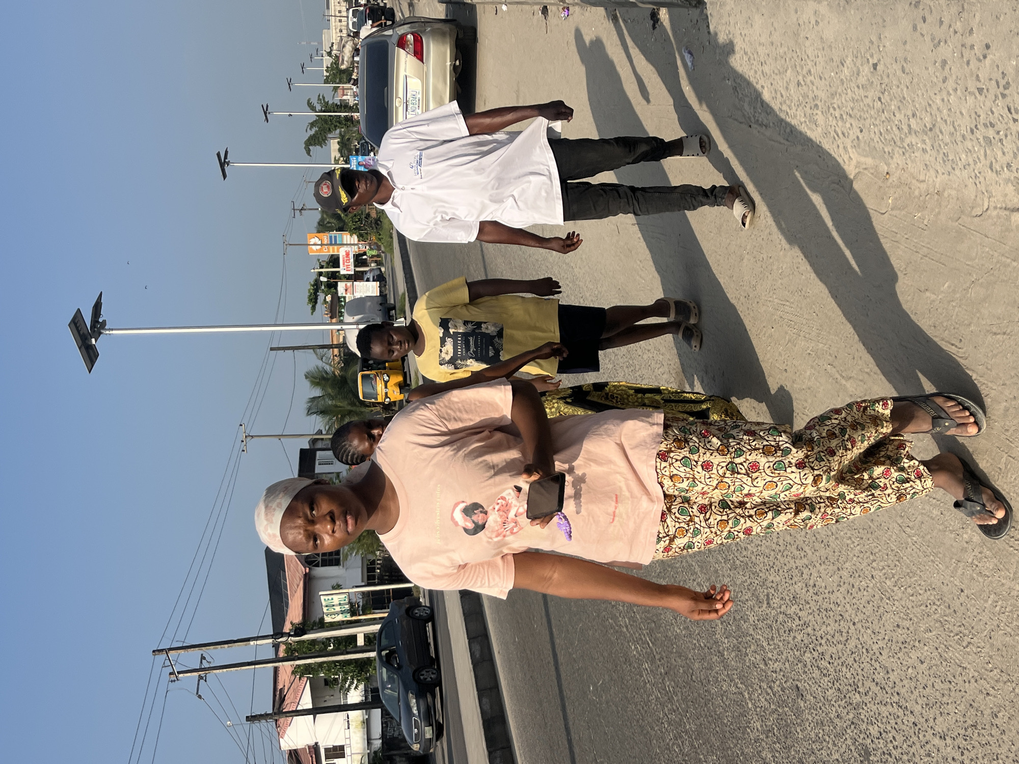The image depicts a street scene in Ado, Nigeria, likely in the early evening due to the long shadows cast by the subjects and the solar-powered streetlights lining the road. Four individuals are walking on a paved sidewalk next to a moderately busy road.
In the foreground, a woman wearing a light pink t-shirt with a graphic print and patterned trousers walks towards the camera, holding a dark-colored mobile phone in her right hand. She has a light-colored head covering and is wearing black flip-flops. Behind her, a woman with braided hair, wearing a yellow t-shirt and dark shorts, walks in the same direction. To the right of these two women, a man wearing a white polo shirt, dark trousers, and a baseball cap is walking. His polo shirt has a logo on the left chest. He is also wearing light-colored sandals. Further behind, partially obscured, is another individual.
The environment includes a paved road with at least one car visible, a concrete sidewalk, and overhead power lines. Buildings with signs, including one for "Dove Hospital," are visible in the background, as are several tall solar-powered streetlights. A yellow tricycle-like vehicle is also present on the road. The sky is clear and blue, indicating fair weather. The overall scene is one of everyday urban life in Ado, Nigeria, with people navigating their environment on foot.