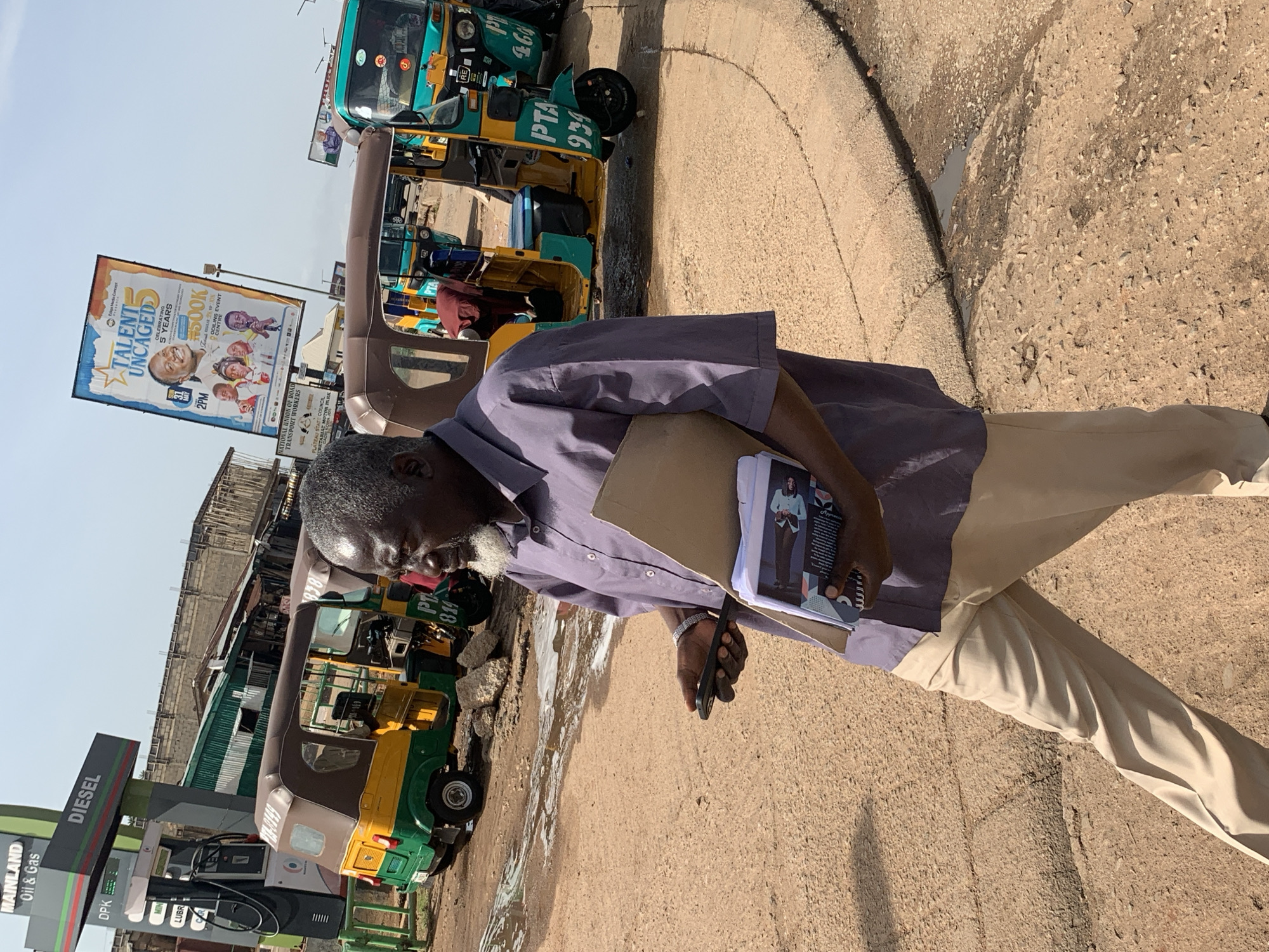 The image depicts a street scene in Gura Top, Nigeria, dominated by a man in a purple shirt and beige pants walking with a phone and a folder. He is holding a dark-colored smartphone in his left hand and a folder with papers in his right hand. In the background, several tricycles, commonly known as 'Okadas' or 'Achaba', are parked. A gas station with "Mainland Oil & Gas" and "Diesel" signage is visible on the left, with a fuel pump. A large billboard advertising "Talent Uncaged 5" is also present in the background. The ground is dusty with some scattered debris, and there are patches of water on the road. The overall impression is a bustling street with local transportation and commercial activities.