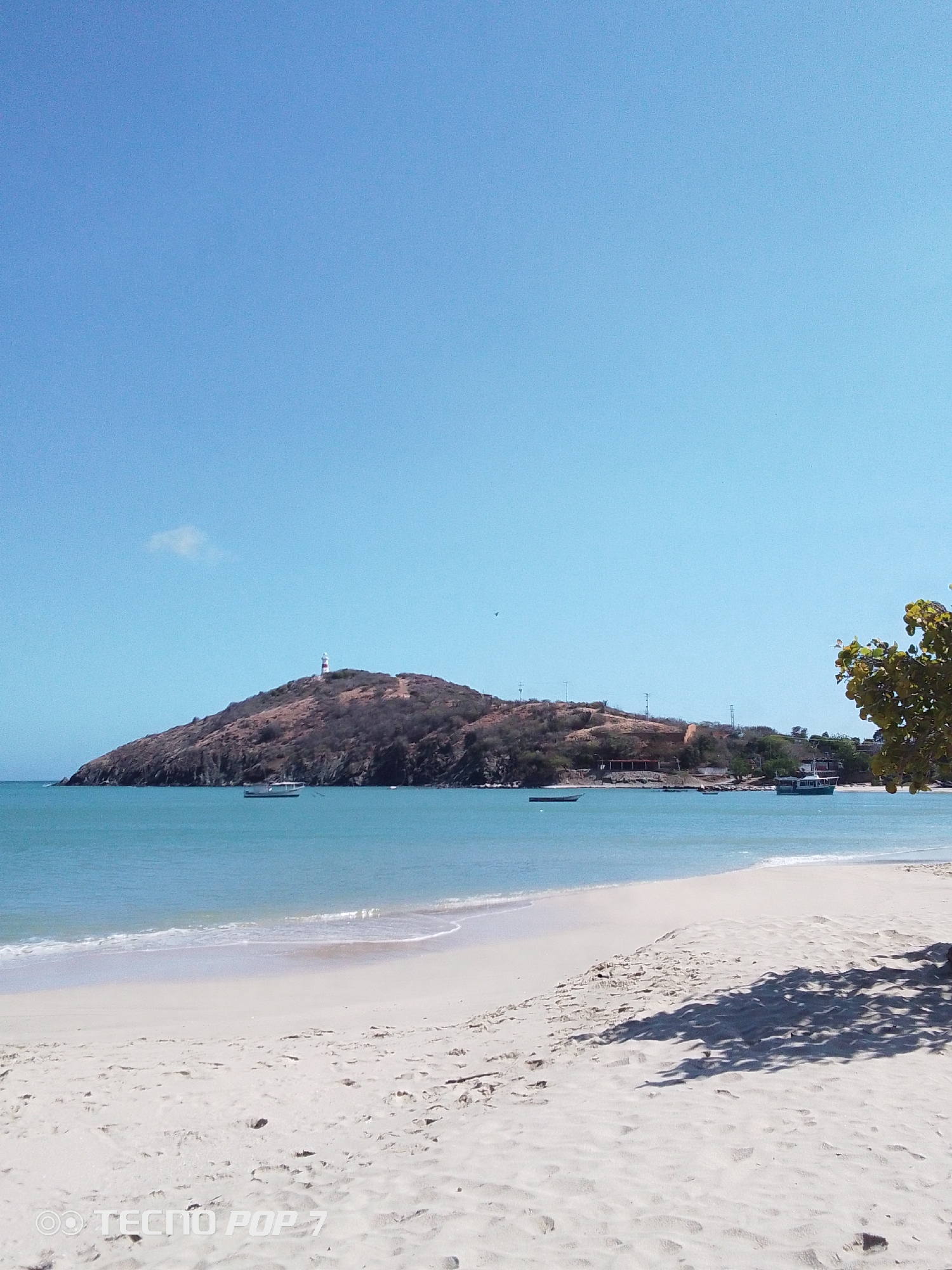 The image depicts a beach scene in Pedro González, Venezuela. The sky is clear and blue. The beach has white sand and gentle waves are washing ashore. In the background, there is a hilly landmass with a lighthouse on top. Several boats are visible on the water near the landmass. There are tree shadows on the sand in the foreground. A "Tecno Pop 7" watermark is present in the lower left corner.