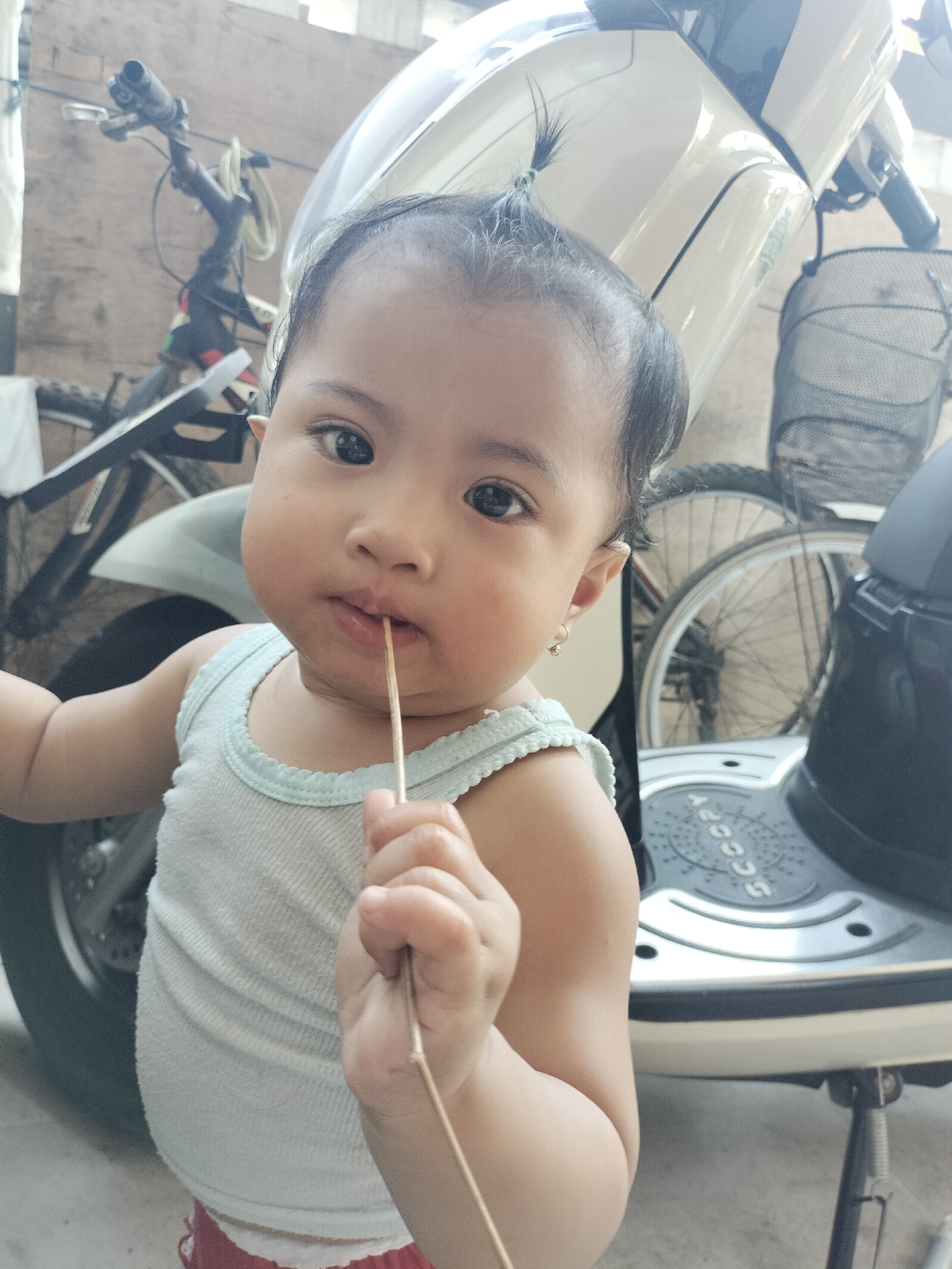In Leces, Indonesia, a close-up, eye-level, medium shot captures a young child holding a thin stick to their mouth. The child, wearing a light blue tank top and red bottoms, has dark hair styled into a small bun with a green tie. Their gaze is directed towards the camera, with one hand holding the stick to their lips. Behind the child, a cream-colored scooter is partially visible, along with the wheel and handlebars of a bicycle. The background appears to be an outdoor setting, possibly a yard or driveway, with a wooden wall.
