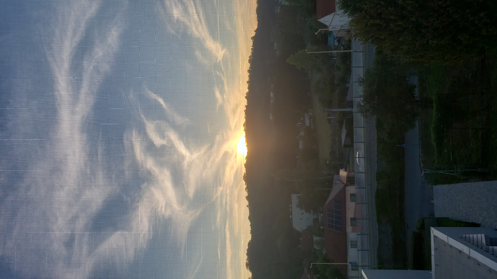 The image depicts a sunset viewed through a window screen, obscuring the view of a suburban landscape in Prešov, Slovakia. The sky is a gradient of blue and orange, with wispy white clouds stretching across. The sun is partially obscured by a tree-covered hill in the middle ground, casting a warm glow over the scene. Several buildings, including houses with solar panels and a larger white building, are visible on the hillside and in the foreground. A street runs horizontally through the mid-ground, with a white car on it. The foreground shows a paved walkway and the corner of a building.