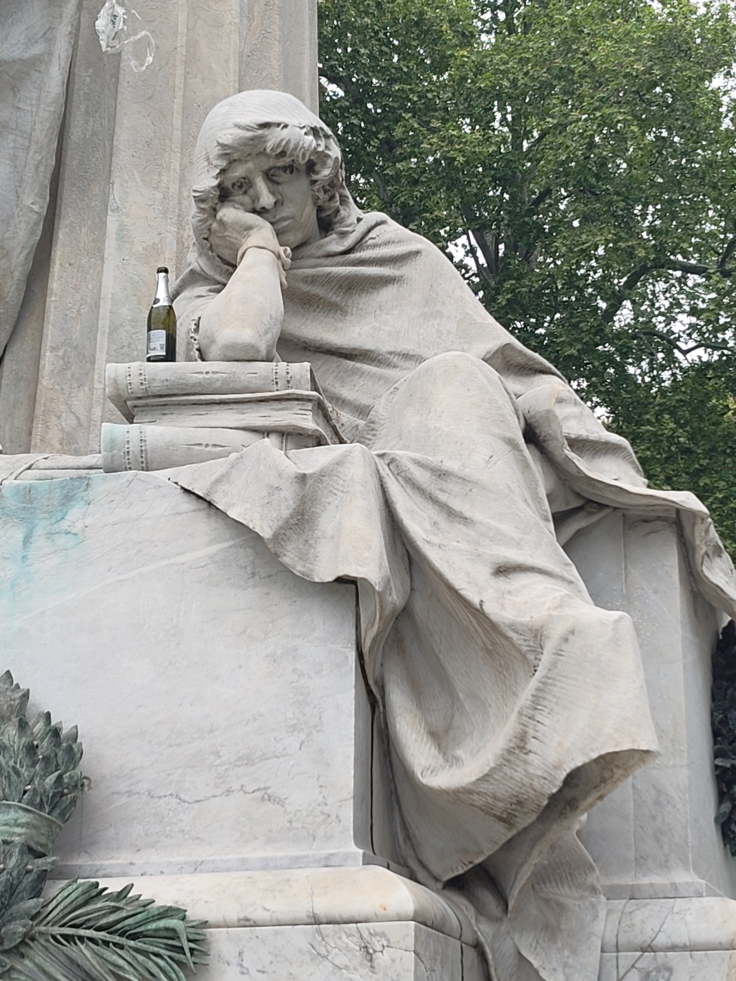 The image depicts a weathered marble statue of a pensive man in a hooded cloak, likely a representation of melancholy or deep thought. He rests his head on his hand, propped up by a stack of books. An open bottle of what appears to be sparkling wine sits atop the books. The statue is part of a larger monument, indicated by the other marble structure to the left and foliage to the right. The scene is lit by diffuse daylight, suggesting an overcast day. This monument is located in the Monumental Cemetery of Staglieno in Genoa, Italy.