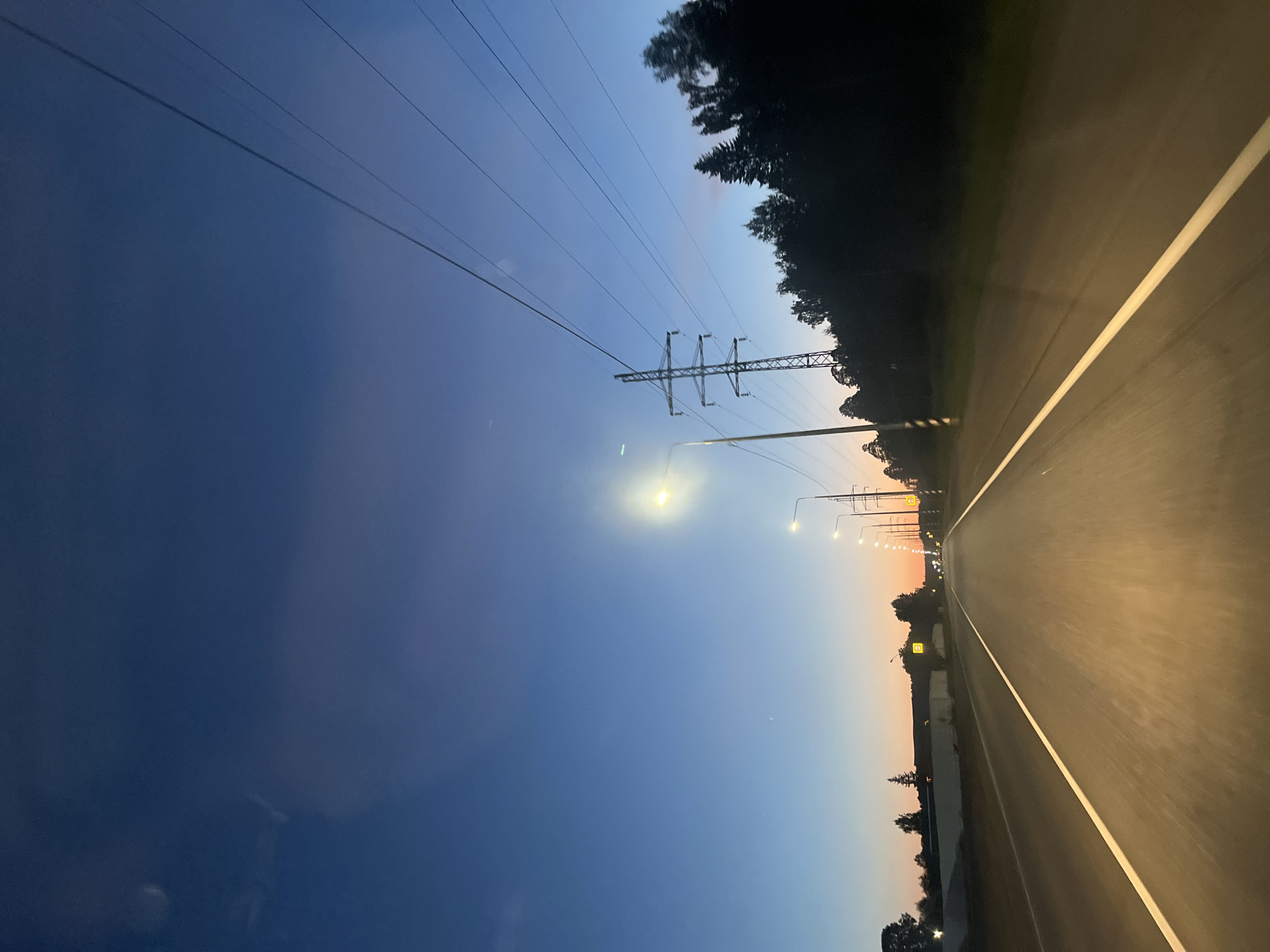 The image, appearing to be captured from a moving vehicle, presents a twilight scene on a road in Chaikovsky, Russia. The perspective looks forward down a long stretch of asphalt highway, likely at dusk or dawn.
The sky dominates the upper half of the frame, showcasing a dramatic gradient of colors. It transitions from a deep, dark indigo blue in the upper left to a lighter azure blue, eventually fading into warm orange and pink hues near the horizon on the right. This coloration strongly suggests either sunset or sunrise.
The asphalt road surface shows signs of motion blur, indicating the vehicle from which the photo was taken is in motion. A distinct, bright white lane marking runs along the right side of the road, guiding the viewer's eye into the distance. Numerous streetlights, emitting intense yellow-white light, illuminate the road ahead and extend into the vanishing point. Some of these streetlights are mounted on poles that also bear small, yellow square signs; these signs appear to have symbols but no legible text is visible. The bright glow of the streetlights creates noticeable halos against the dimming sky.
Along the right side of the road, a dense line of trees forms a dark silhouette against the colorful twilight sky, indicating a forested or rural roadside environment. A prominent metal electricity pylon, characteristic of high-tension power lines, stands tall near the center-right of the image, its lattice structure visible against the sky. Multiple strands of power lines stretch across the upper part of the frame. No people or other vehicles are distinctly visible in the scene. The overall impression is one of peaceful transit during the transitional hours between day and night.