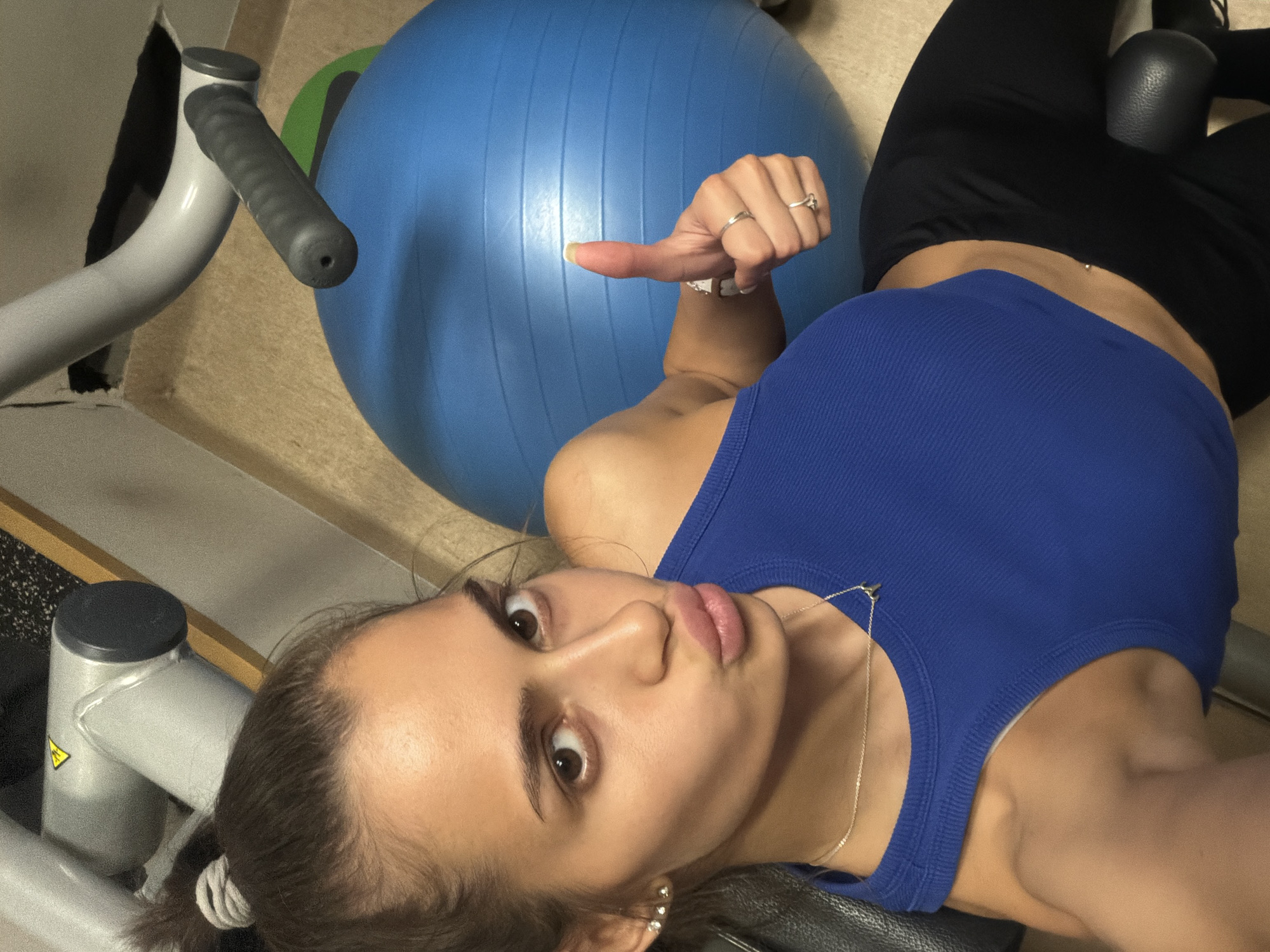 A woman is lying on her back at a slight angle, possibly on a weight bench or similar gym equipment. She has dark hair pulled back and is wearing a blue tank top and black leggings. Her gaze is directed towards the camera, and she's making a slight pouting expression. Her left hand is raised, pointing towards a large blue exercise ball positioned behind her. The surrounding environment seems to be a gym, indicated by the presence of gym equipment like a silver weight machine and other unidentified equipment. The location in the city is Unknown, Unknown.