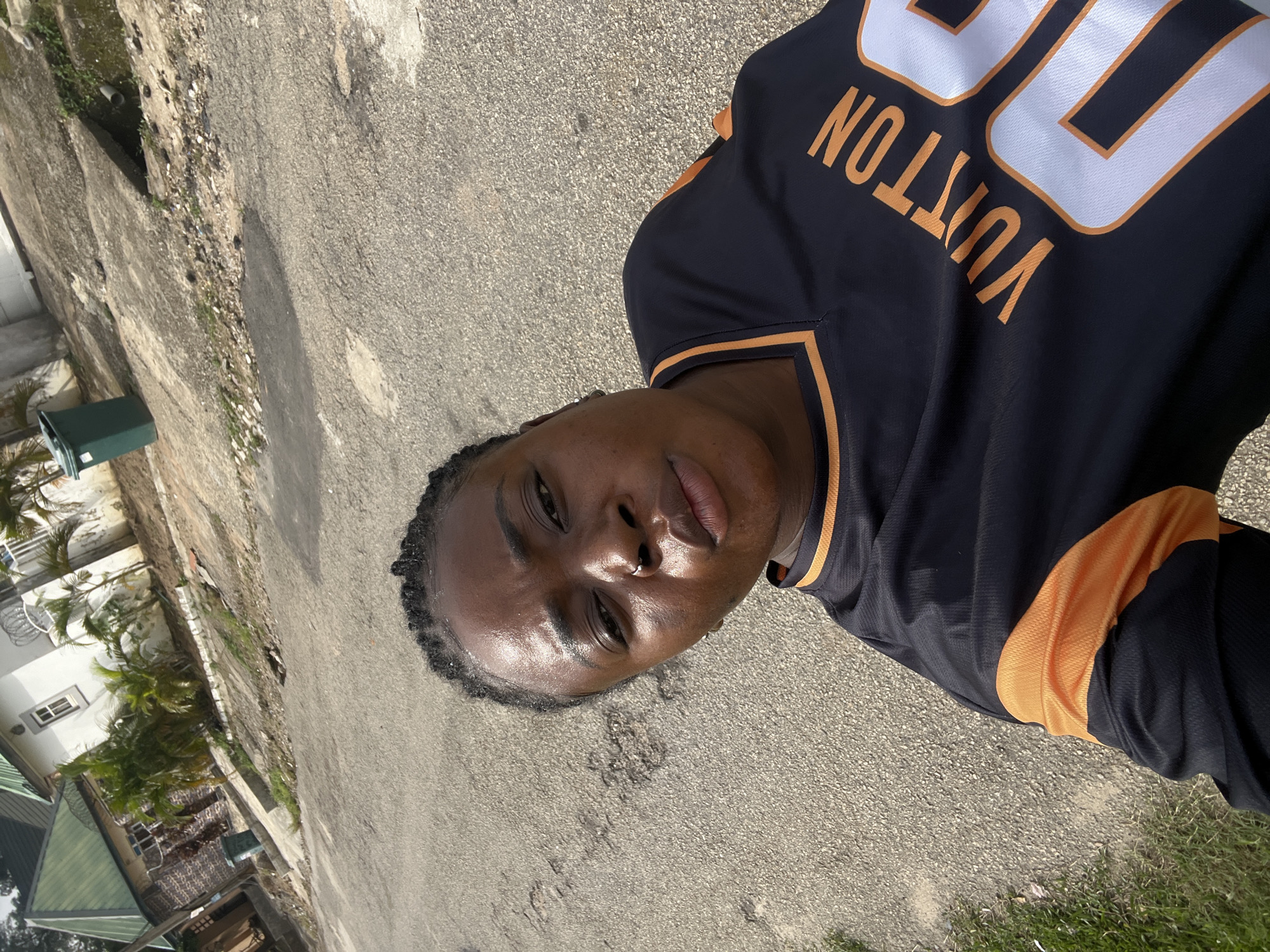 The image is a selfie taken outdoors in an urban environment, likely in Abuja, Nigeria. A young Black woman with dark braided hair and a nose ring is the central subject, wearing a black jersey with orange trim and the word "VUITTON" in orange lettering across the chest. Behind her, the scene includes a paved road with some potholes, a sidewalk, and a residential building with a green roof. A green trash bin is visible near the sidewalk. The ground is a mix of asphalt and dirt with some patches of grass. The lighting suggests it is daytime.
