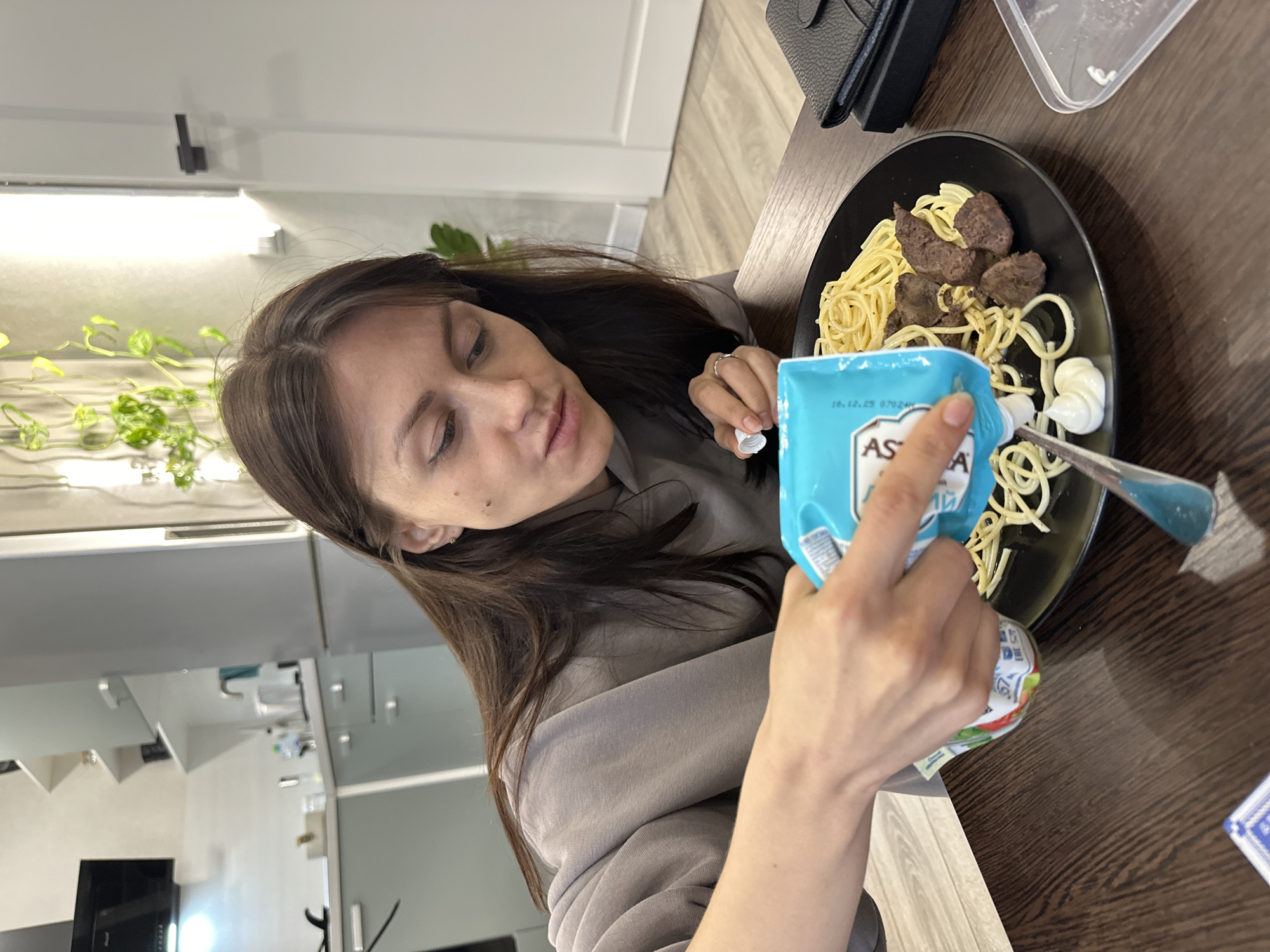 The image captures a woman in an indoor setting, likely a kitchen or dining area in Murino, Russia, focused on preparing or condimenting her meal. She has long, dark brown hair and is dressed in a grey long-sleeved top.
She is actively squeezing a light blue pouch, clearly labeled "ASTORIA" and "МАЙОНЕЗ" (Mayonnaise) in Russian, onto a black plate. A visible date "18.12.23" is printed on the pouch. The plate holds a portion of spaghetti alongside several pieces of dark cooked meat. A silver spoon is positioned to catch some of the white creamy sauce being dispensed.
The table features a dark wooden surface, where a black wallet or small bag and part of a clear plastic container are also visible. The background reveals a modern interior with white walls, a wall-mounted light fixture, and several potted green vining plants on a shelf or ledge. Reflections of kitchen elements, such as cabinets or appliances, can be seen, indicating a contemporary home environment. The woman's expression is one of neutral concentration as she handles her food.