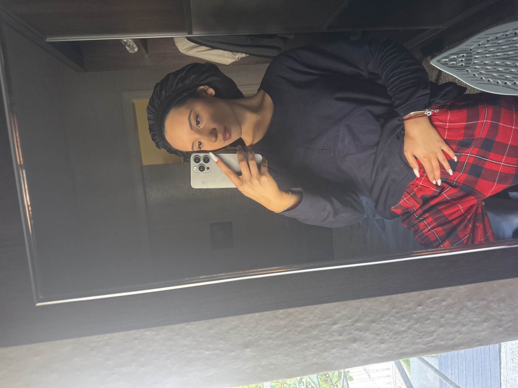 An indoor mirror selfie captures a young adult female in a bedroom or dressing area in Unknown, Unknown. The subject, with medium skin tone and dark hair covered by a black satin bonnet, wears a long-sleeved navy blue t-shirt with a chest pocket and red and black plaid pajama pants. She has white-polished nails, a thin silver bracelet on her left wrist, and a red string bracelet with a silver charm on her right wrist. She holds a light-colored smartphone with her left hand, positioned to take the photo, while her right hand rests on her hip. Her gaze is directed slightly to her left.
The reflection in the mirror shows a dark-framed mirror. To the right of the subject, a dark wooden wardrobe or closet is visible, containing a white shirt and a dark garment. To the left of the mirror, outside the frame, a textured light-colored wall is present, and through a window or glass door, green foliage and an outdoor balcony with decking and railings are visible. The bottom right of the image shows the silver-grey base of a clothing iron and a portion of a patterned rug or carpet. Natural light illuminates the scene, seemingly from the direction of the outdoor area.