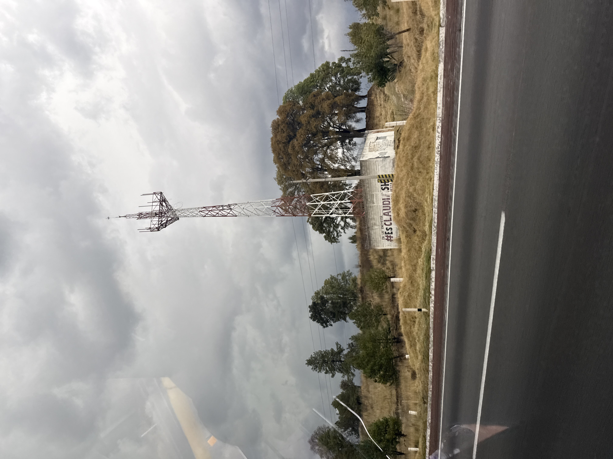The image shows a roadside view near Santa Rita Tlahuapan, Mexico. A red and white telecommunications tower stands in a field adjacent to a highway. Next to the tower is a wall or building covered with graffiti, including the hashtag "#ESCLAUDIA". The landscape consists of dry grasses and trees, with a cloudy sky above. The highway has a black asphalt surface with a white dividing line. The image appears to be taken from inside a vehicle, with reflections visible on the windshield.