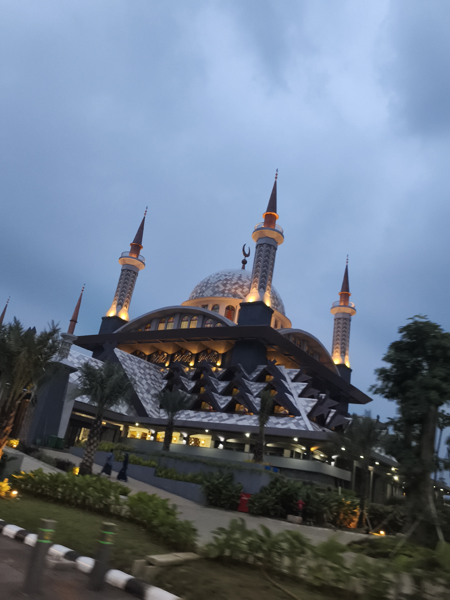 This image depicts a striking, modern mosque at dusk or early night, located in Cibinong, Indonesia. The sky is a deep, cloudy blue-grey, providing a dramatic backdrop for the illuminated structure.
The mosque features a prominent central dome adorned with intricate patterns and topped with a crescent moon finial. Multiple slender minarets, each with a pointed spire and illuminated band, rise from different points of the mosque. The building's main body showcases a complex, multi-tiered roof system composed of numerous interconnected geometric or triangular facets, also displaying elaborate patterns. Warm, golden light spills from within the mosque and illuminates its exterior details, highlighting its unique architectural style.
In the foreground and surrounding the mosque is lush green landscaping, including palm trees and various bushes. A paved pathway is visible in the lower left, where two figures, appearing to be individuals in traditional long garments, are walking away from the viewer. A black and white striped barrier or curb runs along the very bottom left edge of the frame, suggesting a roadside or designated pathway. The overall scene captures the mosque's grandeur and its contemporary design, beautifully accented by the evening illumination against the twilight sky. No visible text is clearly discernible in the image.