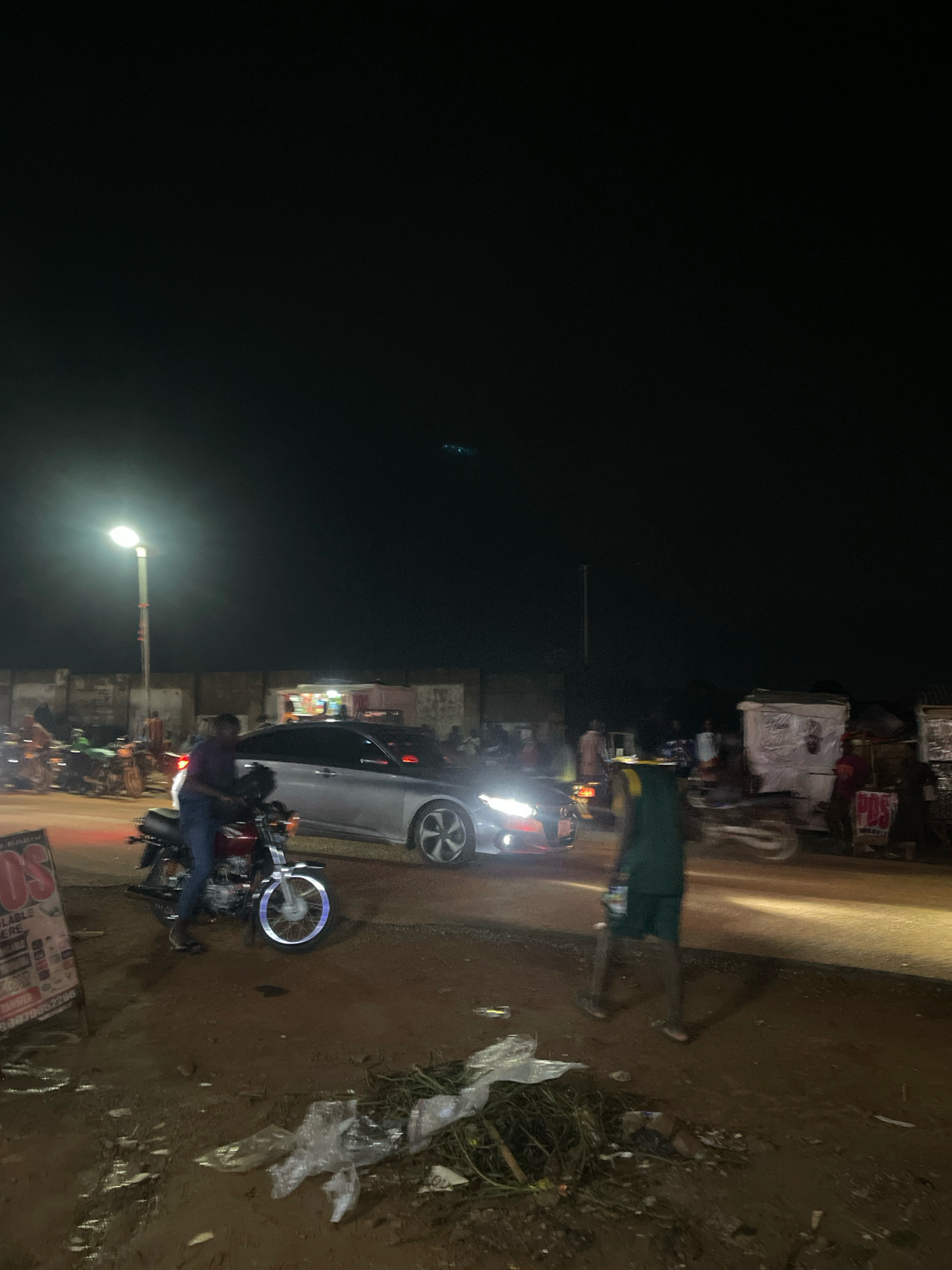 The image depicts a street scene at night in Abuja, Nigeria. A street lamp illuminates a busy intersection where several motorcycles and a car are present. On the left side of the image, a man is sitting on a red motorcycle. A silver car is visible in the middle of the road, with its headlights on. Further down the road, there are some stalls and more people. The ground in the foreground is muddy and littered with debris, including plastic bags and vegetation. The overall impression is of a vibrant, albeit somewhat unkempt, urban night environment.