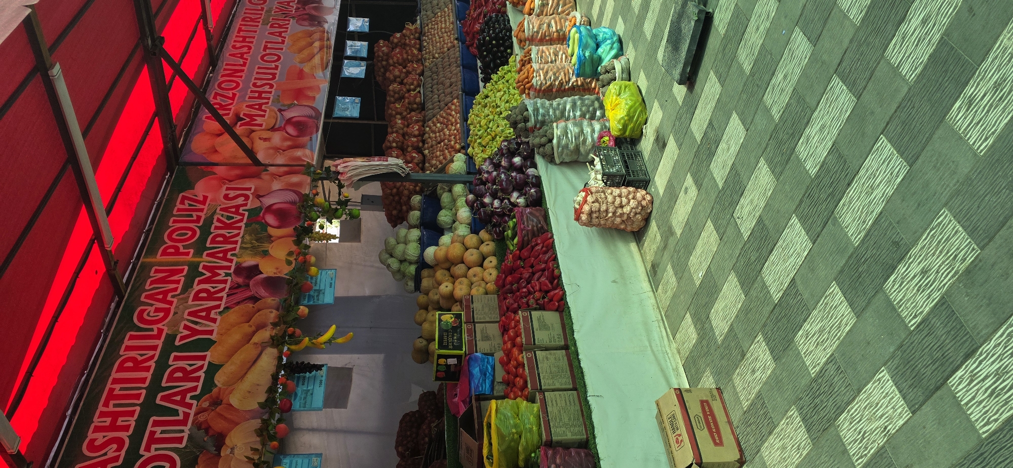 The image shows a market stall in Tashkent, Uzbekistan, selling a variety of fresh produce. The stall is covered by a red awning. A banner above the produce reads "ASHTIRILGAN POLIZ MAHSULOTLARI YARMARKASI" which translates to "Grown Produce Fair."
Various vegetables are displayed, including tomatoes, bell peppers, eggplants, onions, and cabbages. There are also some fruits visible, such as bananas and grapes, arranged decoratively above the vegetables. Some of the produce is displayed in woven bags and crates, while other items are piled high on the stall. A cardboard box with the text "RBO KOKOS YOG'I" meaning "RBO Coconut Oil" is in the foreground. The ground is paved with a patterned tile in shades of green and grey. There are no people visible in the image.