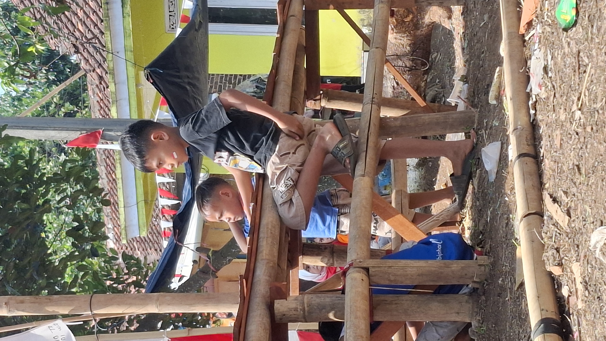 The image captures a scene outdoors in Cilaku, Indonesia, with a focus on two young boys. One boy, wearing a black t-shirt and khaki shorts with a pattern, is seated with his legs crossed on a bamboo structure. He has red and white paint on his face, suggesting participation in a patriotic event or celebration. His gaze is directed to the left. Beside him, another younger boy in a blue t-shirt looks towards the left, away from the camera. The background reveals a yellow building with a tiled roof and trees. Red and white triangular flags are visible, attached to a black fabric canopy, further indicating a festive or nationalistic occasion. The ground is dirt and littered with debris, including plastic cups and wrappers. Bamboo poles form the structure the boys are sitting on, and another bamboo pole lies horizontally in the foreground.