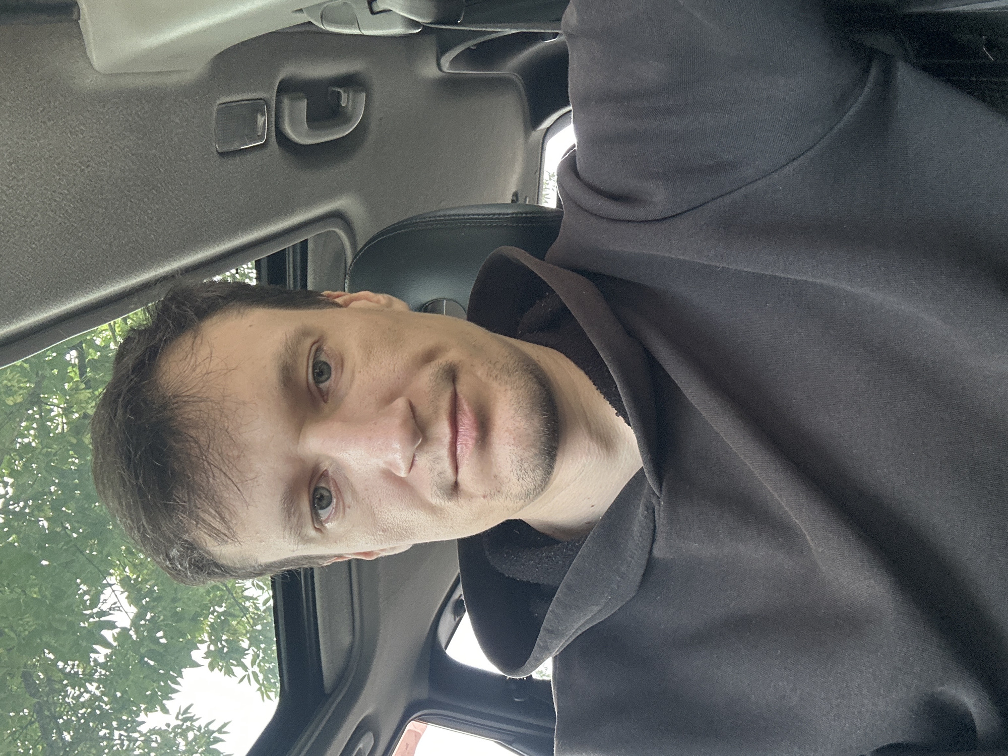 A man with short brown hair and green eyes is seen in a close-up, selfie-style shot from inside a car. He is wearing a dark grey hooded sweatshirt. The car's sunroof is open, revealing green foliage and a cloudy sky above. The interior of the car, including the grey headliner and a black car seat, is visible. The man's facial expression is neutral. The setting appears to be daytime. The image was taken in the city of Makiv, Ukraine.