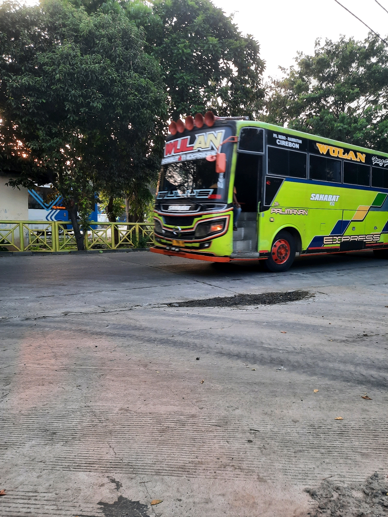 The image shows a green and yellow bus with the brand name "WULAN EXPRESS" written on its side. The bus is parked on a concrete road, possibly at a bus station, with trees and a yellow fence in the background. The bus has its door open, and a person can be seen inside. The scene appears to be in a tropical or subtropical climate, given the lush greenery. The lighting suggests it is either early morning or late afternoon. The bus has several lights on its roof and a driver's cabin with a prominent windshield. The overall atmosphere is that of a transportation hub.