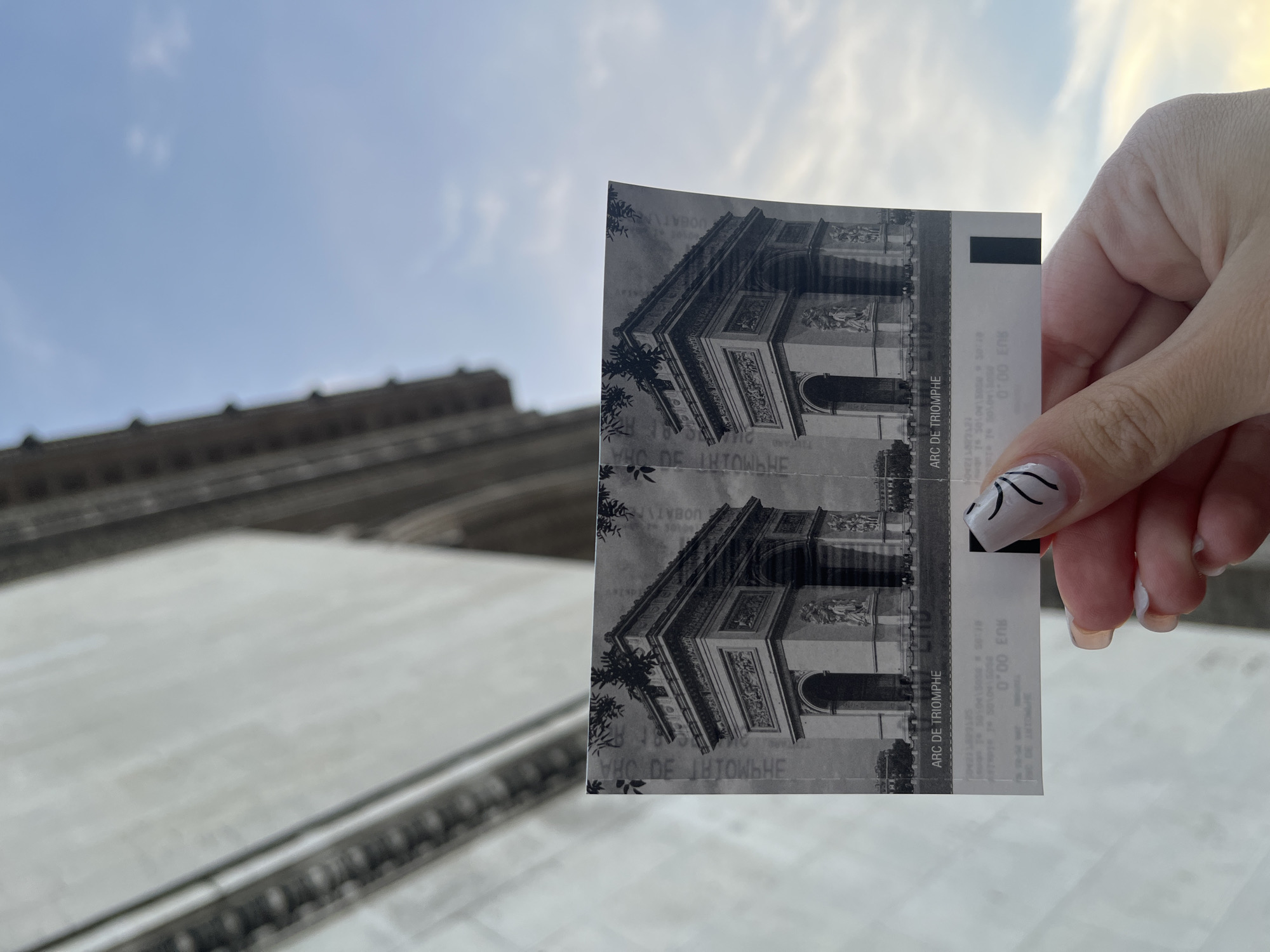 A close-up, slightly upward-angled shot captures a person's right hand holding two black and white tickets against a bright sky. The tickets, appearing stacked, prominently feature an image of the Arc de Triomphe and are clearly labeled "ARC DE TRIOMPHE" multiple times. Other visible text on the tickets includes "0.00 EUR" and partially obscured or reversed elements such as "E JUJOWHE HE", "UOBAT PEDA", and "АНТОШЬНЕ". The hand has manicured nails with a white base and black abstract line art.
The background is softly out of focus, revealing a light blue sky with faint white clouds at the top. Below the sky, the blurry, light-colored stone facade of a large architectural structure is visible, consistent with the majestic scale and material of the Arc de Triomphe itself. This visual context, combined with the explicit text on the tickets, strongly indicates the scene is set at or very near the Arc de Triomphe in Paris, France. The bright lighting suggests it is daytime under clear or partly cloudy skies. The photograph captures a common tourist moment, showcasing visit tickets to a renowned Parisian landmark.