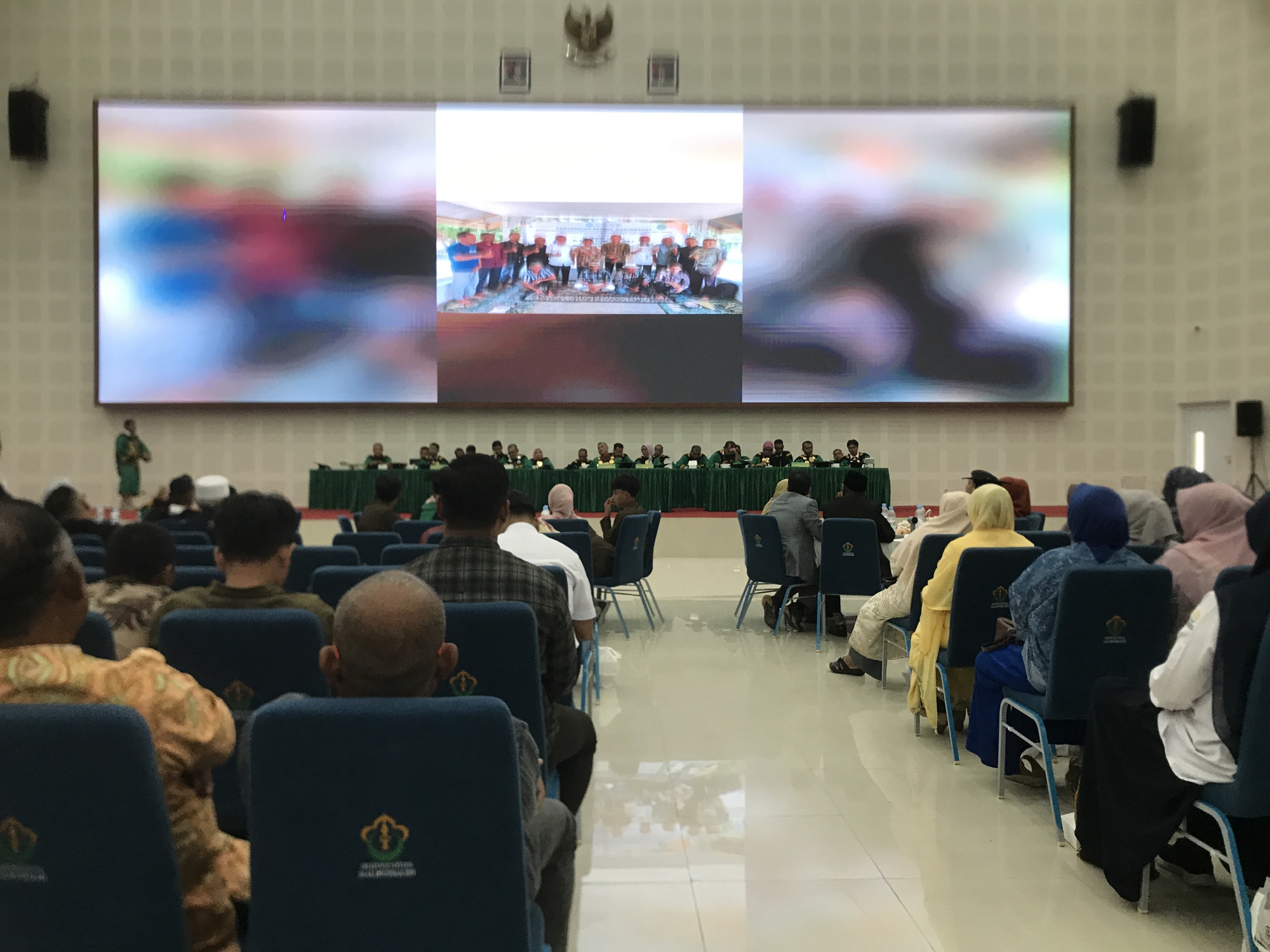 A large auditorium in Reuleut Timu, Indonesia, is filled with seated individuals facing a large screen displaying a group photo on a stage. Many attendees are wearing traditional Indonesian clothing and head coverings. A row of officials sits at a table at the front of the room, and the floor is a polished surface. The screen is flanked by speakers mounted on the wall.