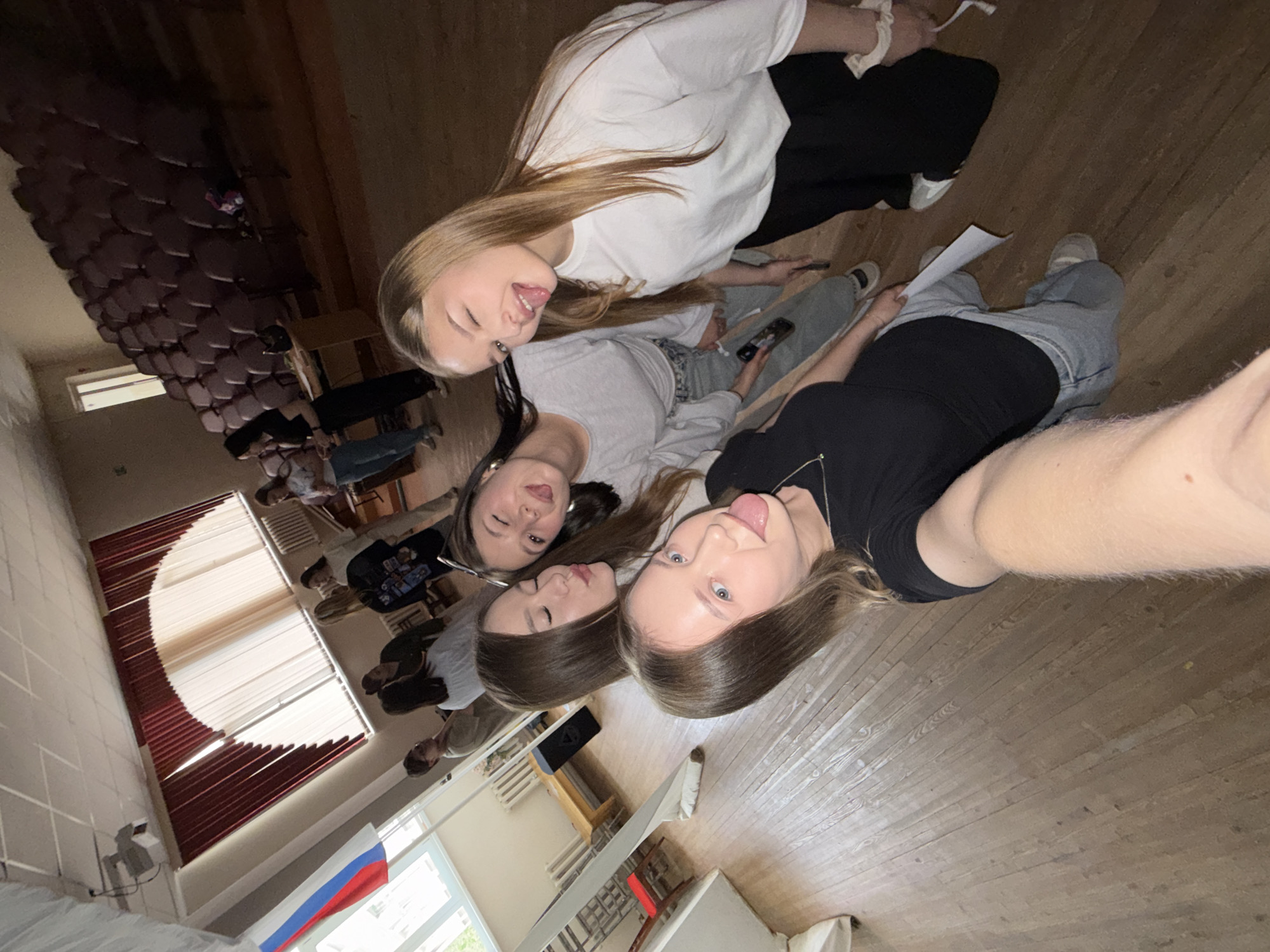 The image shows a selfie taken indoors by one of four young women. The women are posing for the photo, sticking their tongues out. They are dressed in casual clothing. The background shows a classroom or assembly hall with rows of seats. A Russian flag is visible in the lower left corner. The location is most likely a school or similar institution in Chaikovsky, Russia.