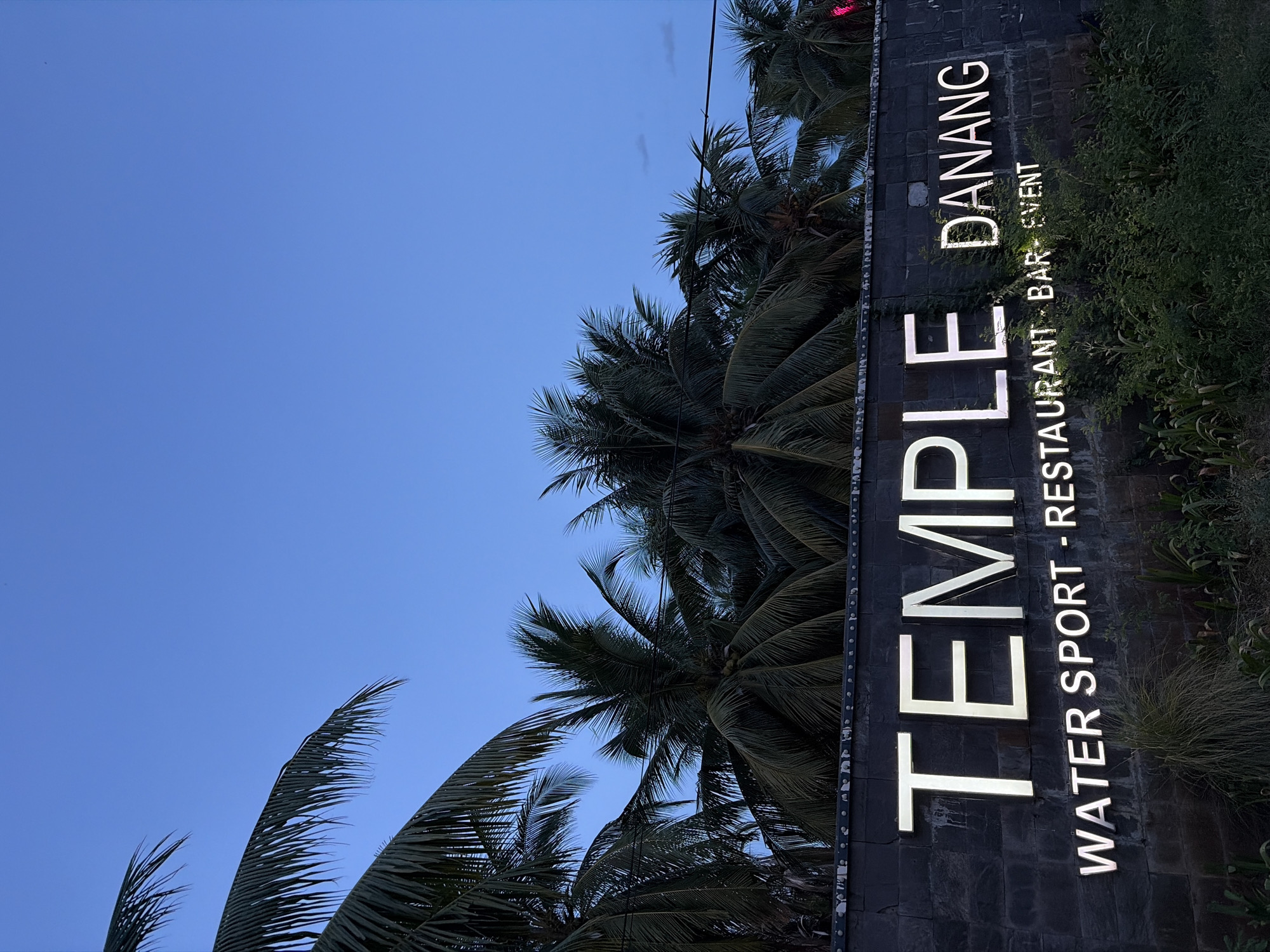 The image shows a large illuminated sign that reads "TEMPLE DANANG WATER SPORT - RESTAURANT - BAR - EVENT" against a backdrop of palm trees and a twilight sky. The sign is mounted on a dark stone wall. The lighting suggests it is either dusk or dawn. The location is identified as Đà Nẵng, Vietnam, a coastal city known for its beaches and water sports.